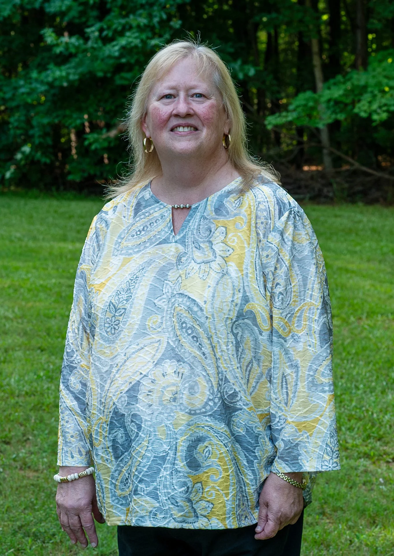 A woman with blonde hair wearing a patterned blouse and jewelry standing outdoors on a grassy area with trees in the background.