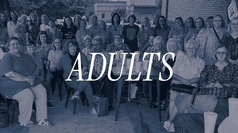 Group of adults sitting and standing outdoors, smiling, with some seated on chairs and others standing behind, during daytime.