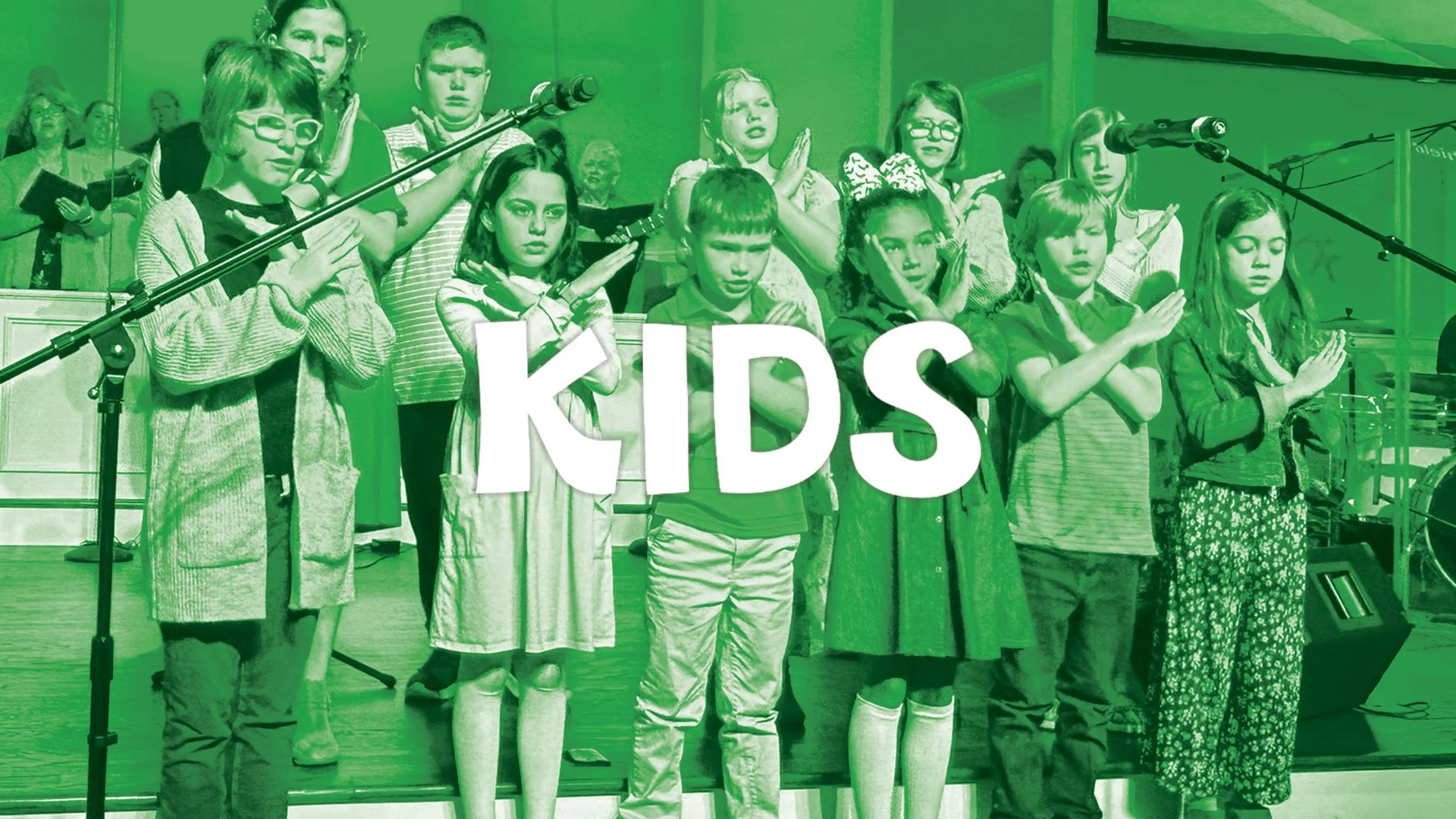 Children standing on stage with hands crossed in front of their chests, participating in a Kids' event or performance, with a woman and other performers in the background.