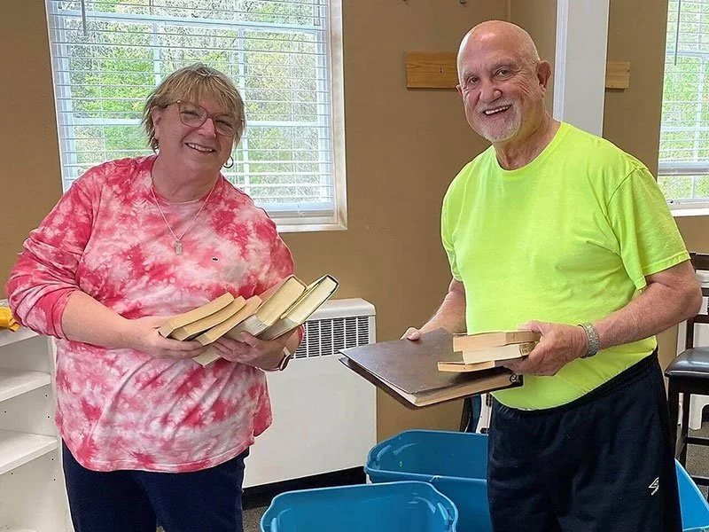 A woman in a pink tie-dye shirt holding books, smiling, standing next to a man in a bright yellow shirt, also holding books, in a room with windows and a blue plastic bin on the floor.