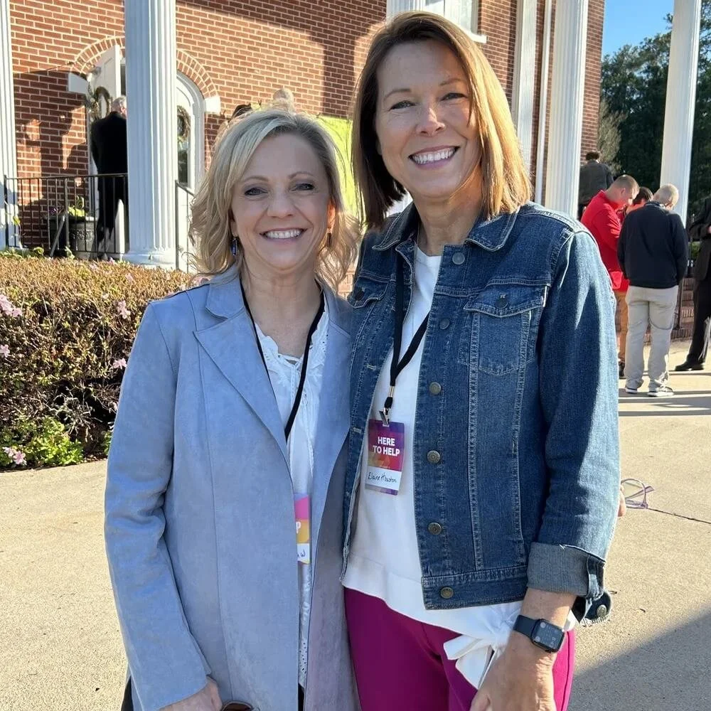 Two women smiling outdoors in front of a red brick building with white columns and greenery, at a sunny event.