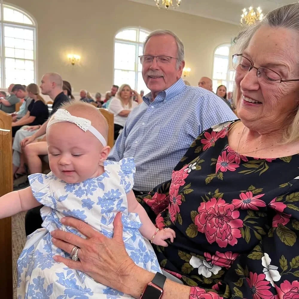 A baby girl wearing a blue floral dress and a white headband, sitting on a woman's lap, with people in the background inside a church or similar venue.
