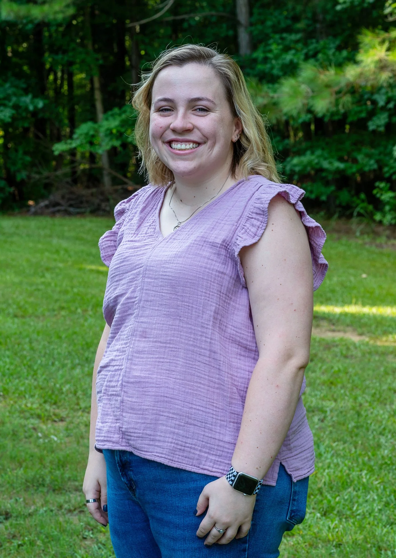 A young woman in a purple sleeveless top stands outdoors on grass, smiling with trees in the background.