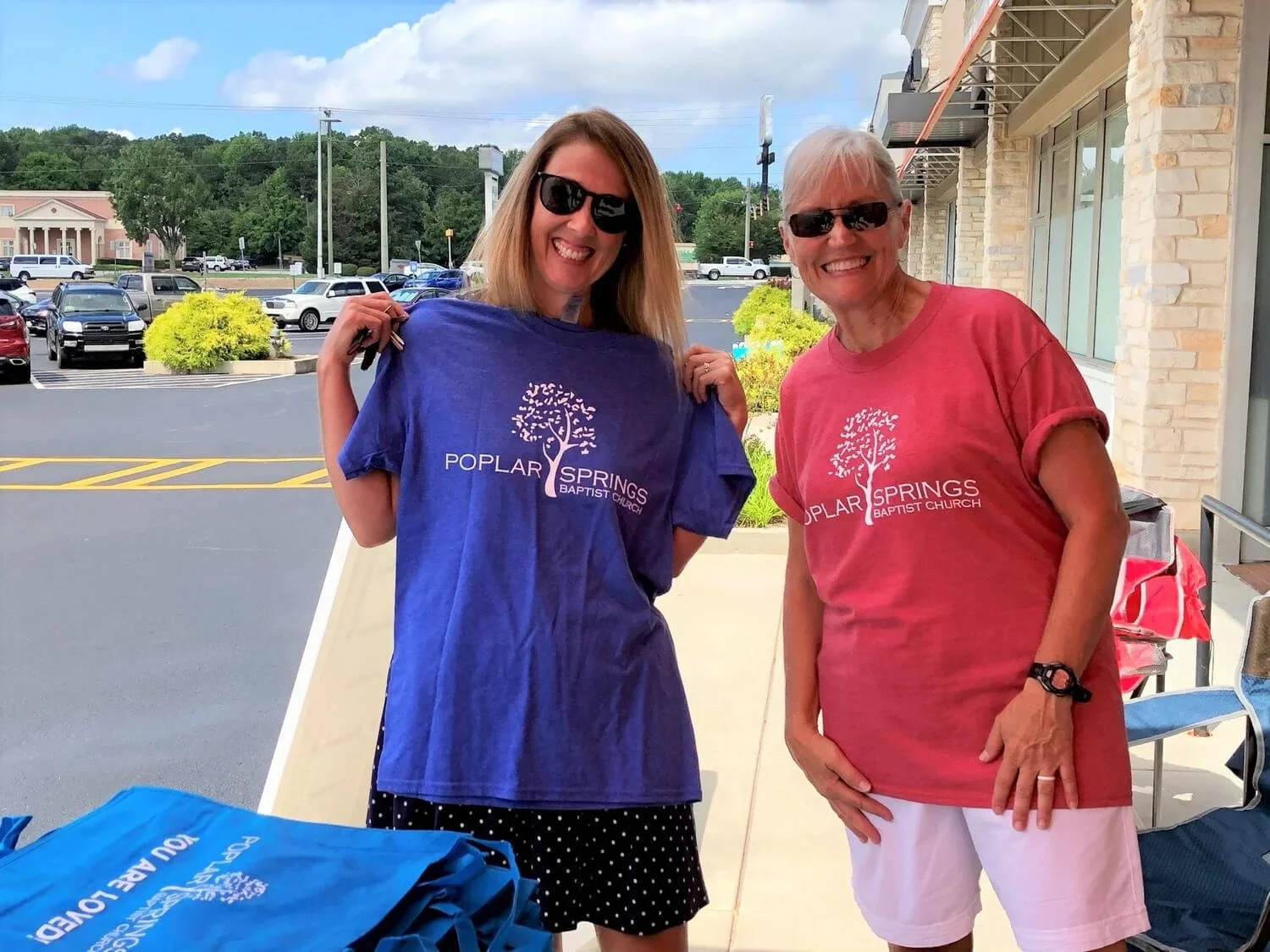Two women smiling outside of a building, holding up T-shirts with the logo of Poplar Springs Baptist Church