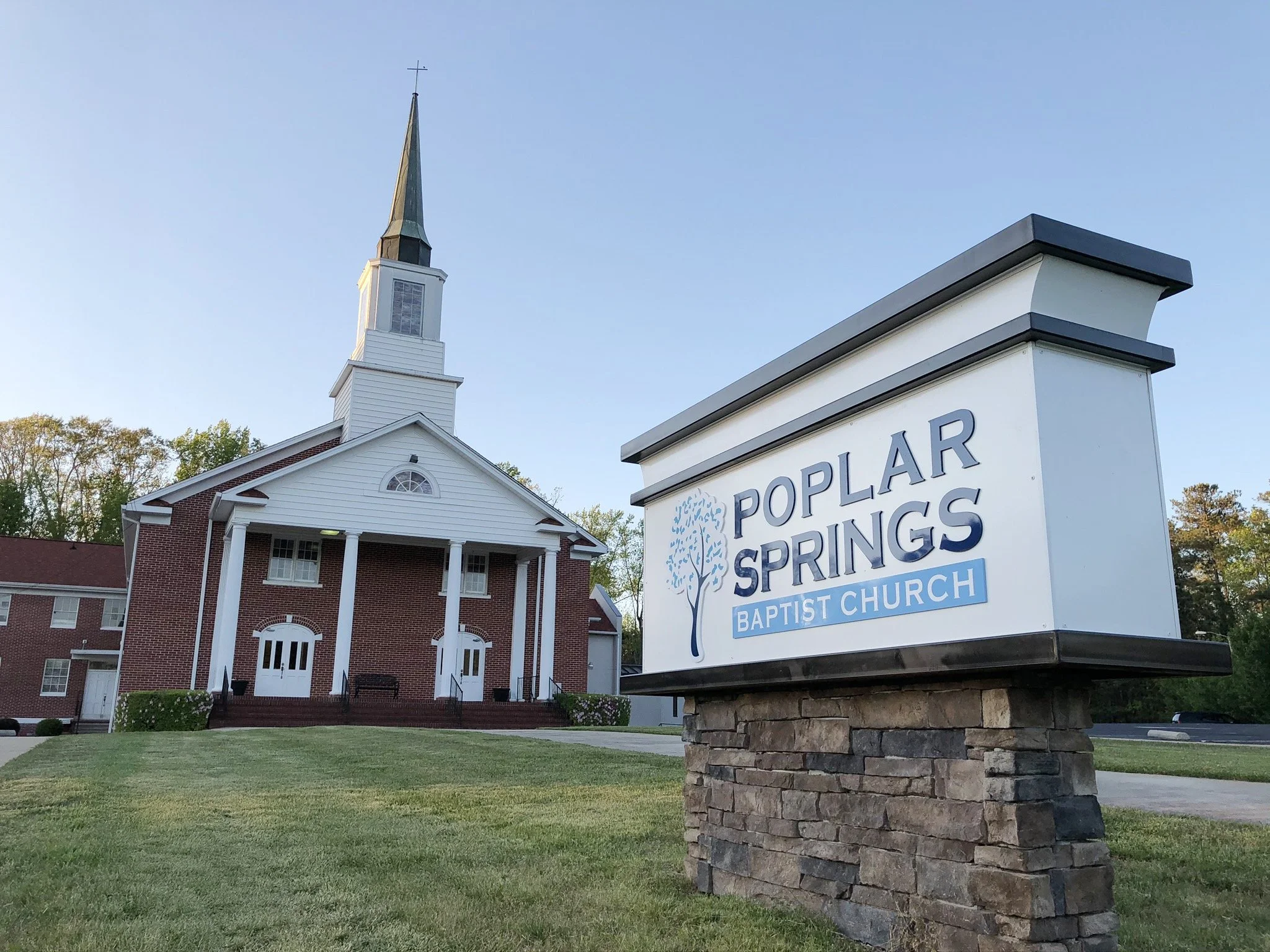 A church building with red brick walls, white columns, and a tall steeple with a cross on top. In the foreground, a sign reading "Poplar Springs Baptist Church" is mounted on a stone base, visible on the lawn of the church.