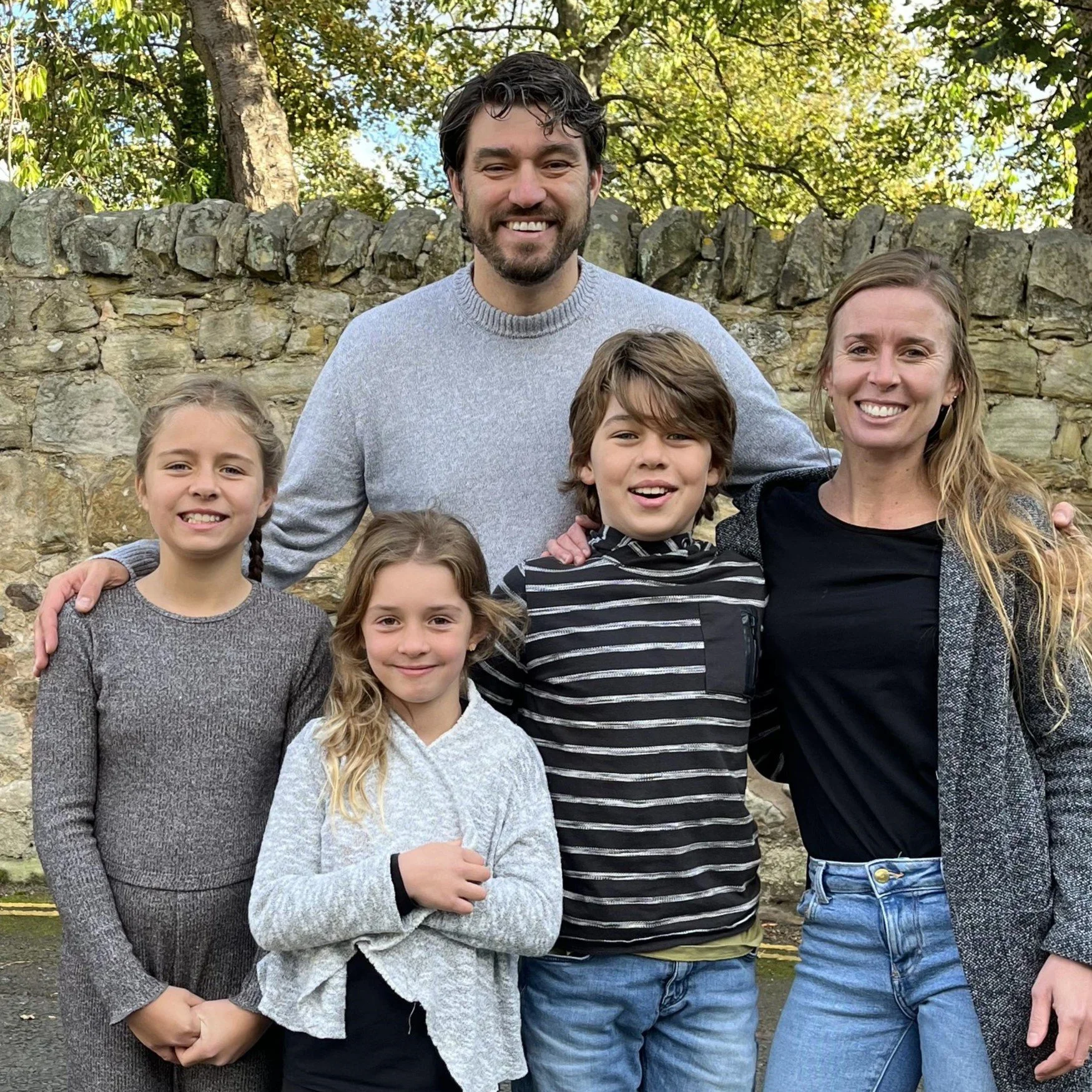 A family of six smiling outdoors in front of a stone wall and trees.