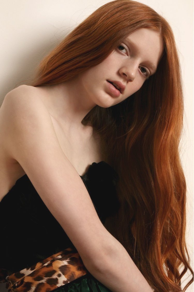 A young woman with long, red hair and fair skin, looking at the camera, wearing a black strapless top and animal print clothing.