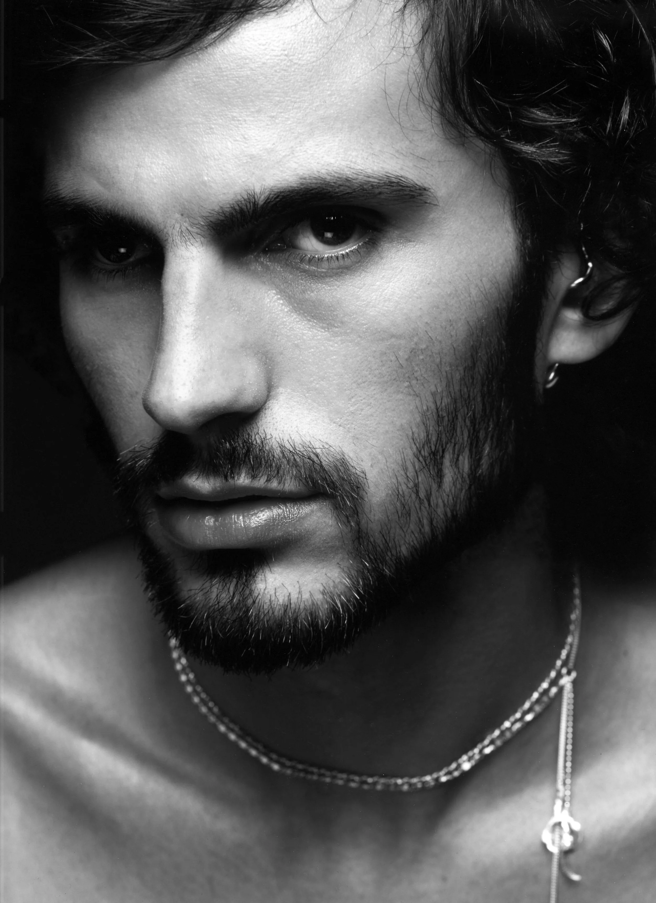 Black and white close-up portrait of a man with dark hair, beard, mustache, earrings, and layered necklaces, looking directly into the camera.