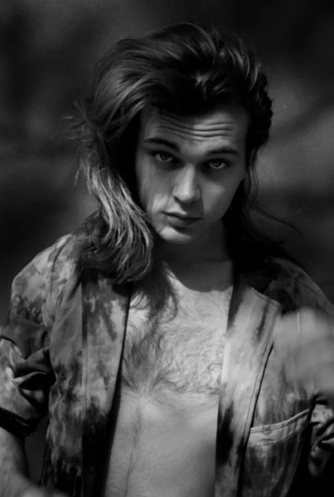 Black and white photo of a young woman with long, wavy hair, wearing a tie-dye shirt and jacket, with a serious expression.