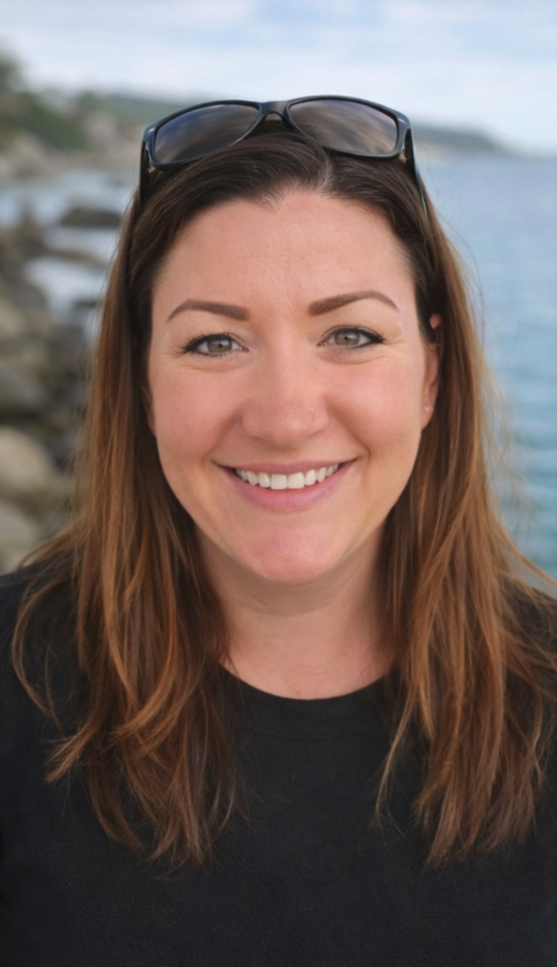 A smiling woman with long brown hair, wearing sunglasses on her head, standing outdoors near a rocky shoreline with water in the background.