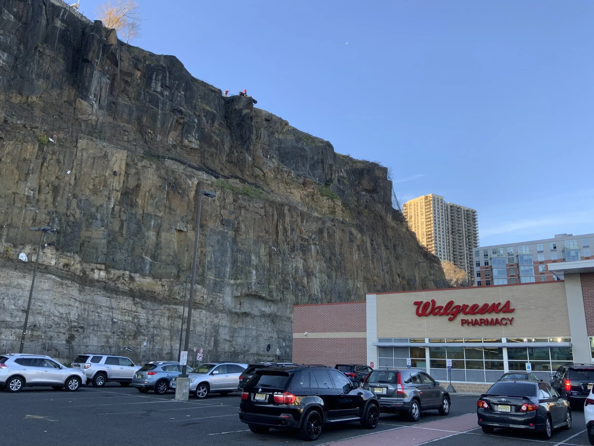 A parking lot in front of a Walgreens pharmacy, with cars parked and a large rocky cliff behind the store. There are some people visible at the top of the cliff and a tall building in the background.