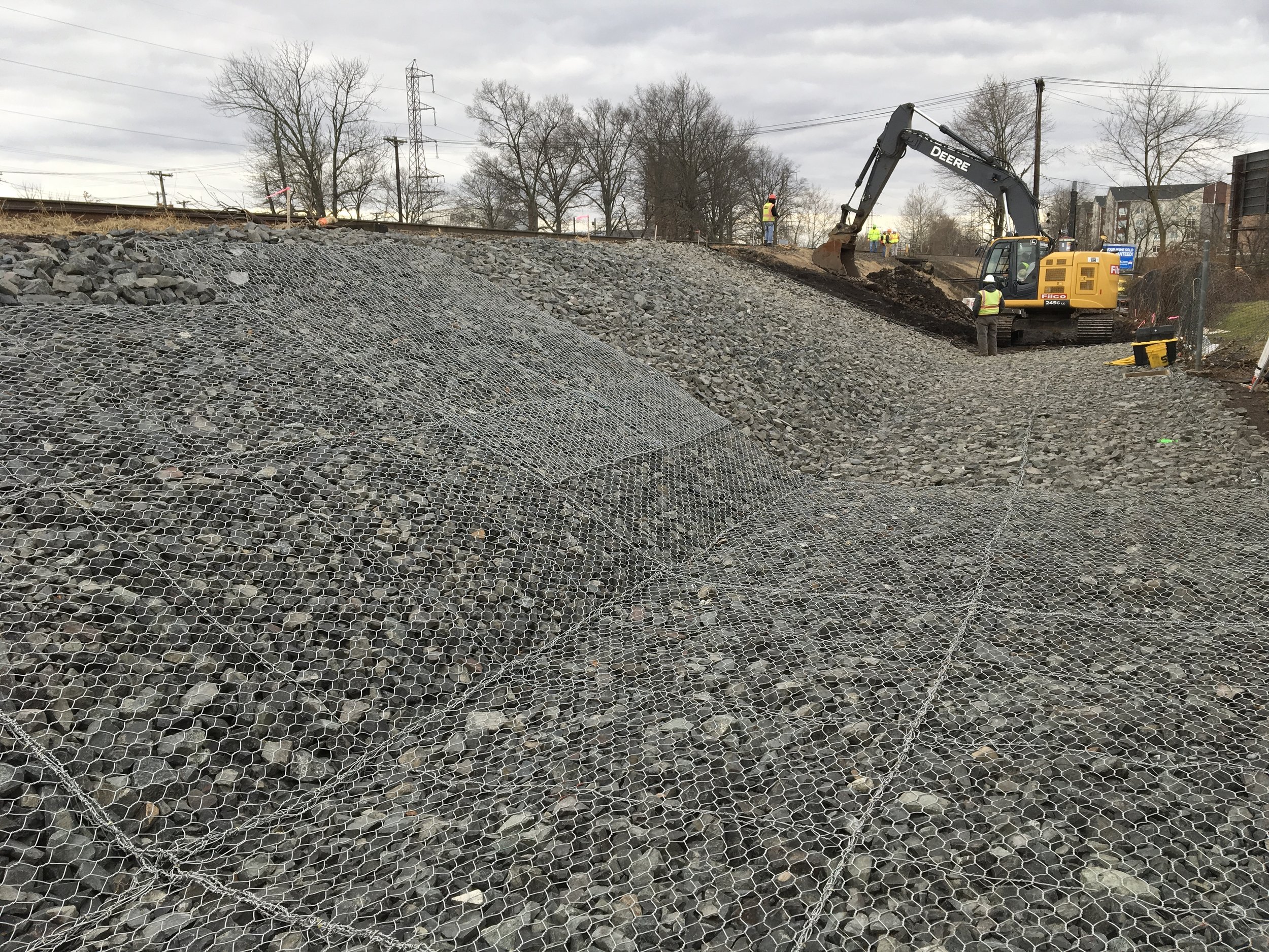 Construction workers and an excavator working on a railway embankment with rock and wire mesh fencing under an overcast sky.