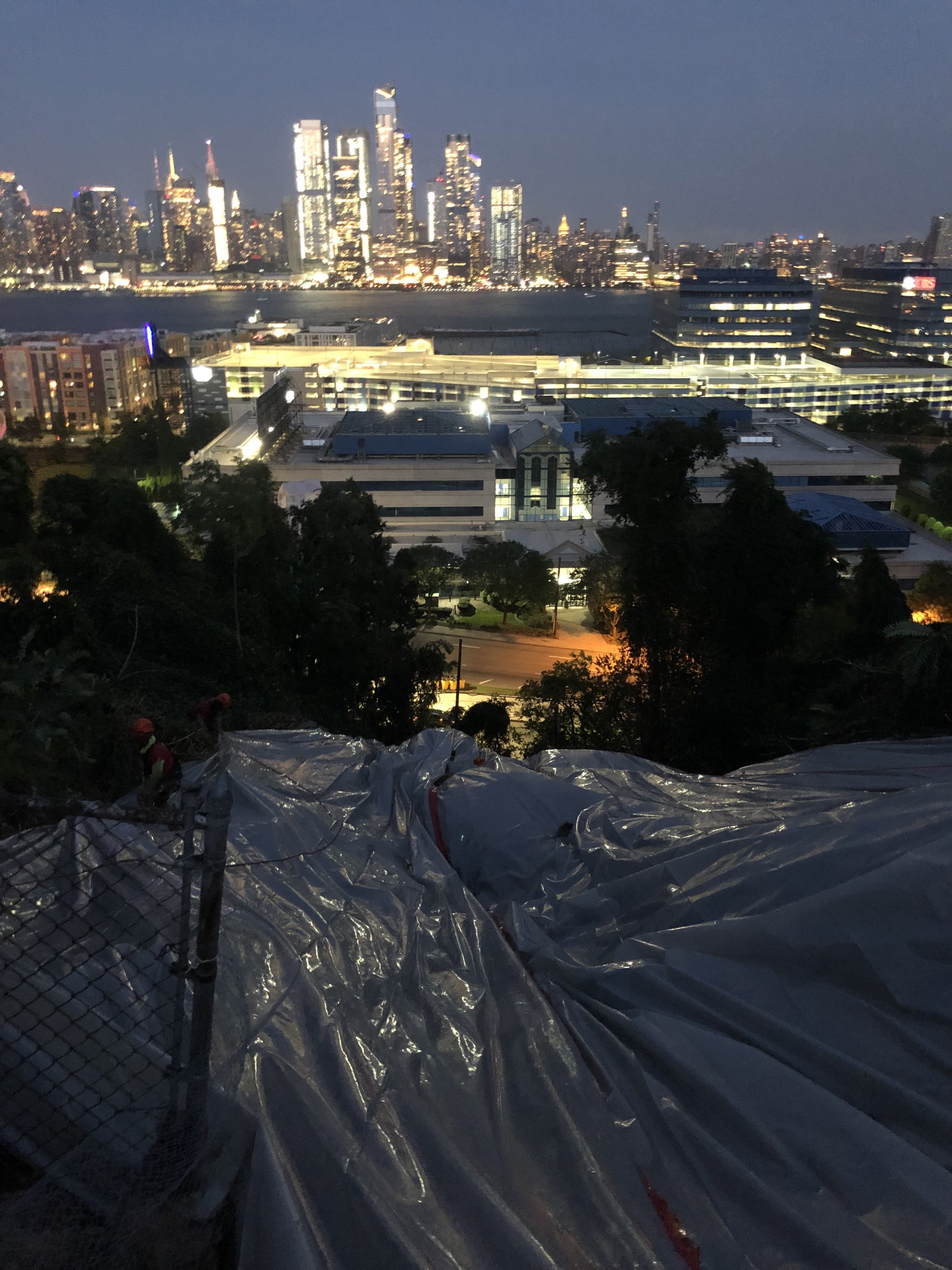 A city skyline at night with tall illuminated buildings across a body of water, view from a hillside with construction workers and plastic sheeting in the foreground.