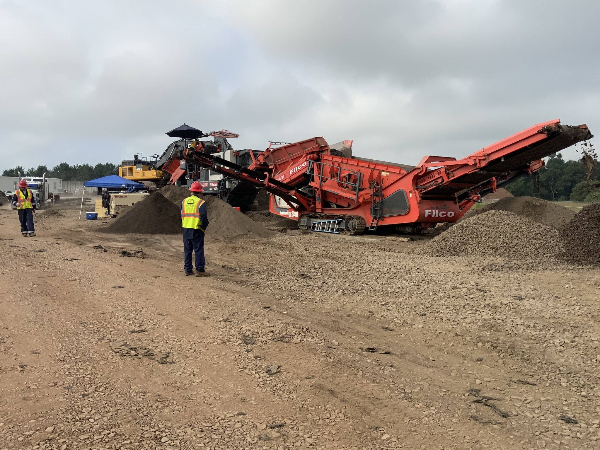 Construction workers operating a large orange asphalt paver on a dirt road, with piles of gravel and a cloudy sky in the background.
