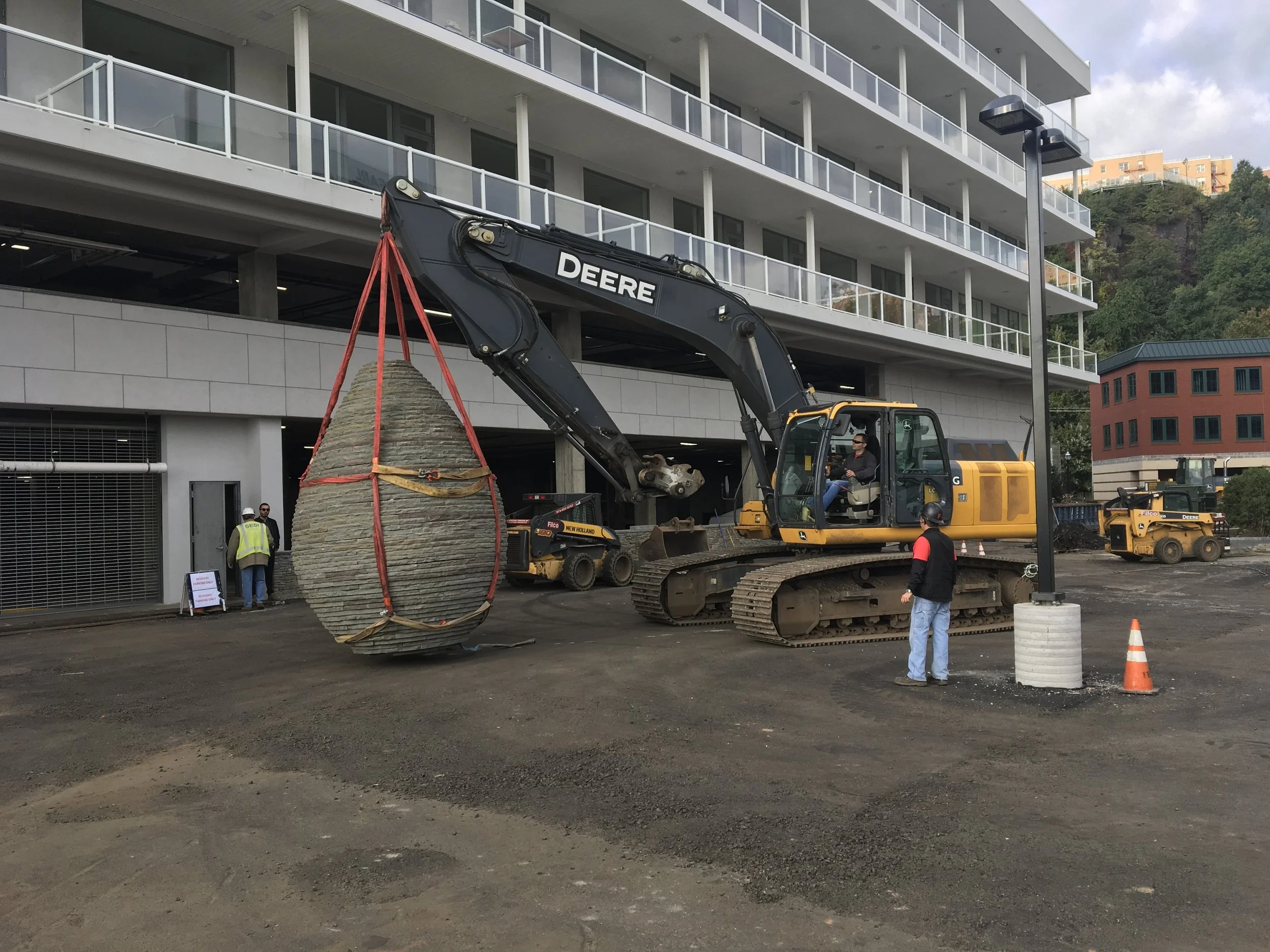 Construction site with a large John Deere excavator lifting a big, circular stone slab with red lifting straps. Several workers, some wearing safety vests and helmets, are present. The background shows a modern building with balconies and a hillside 