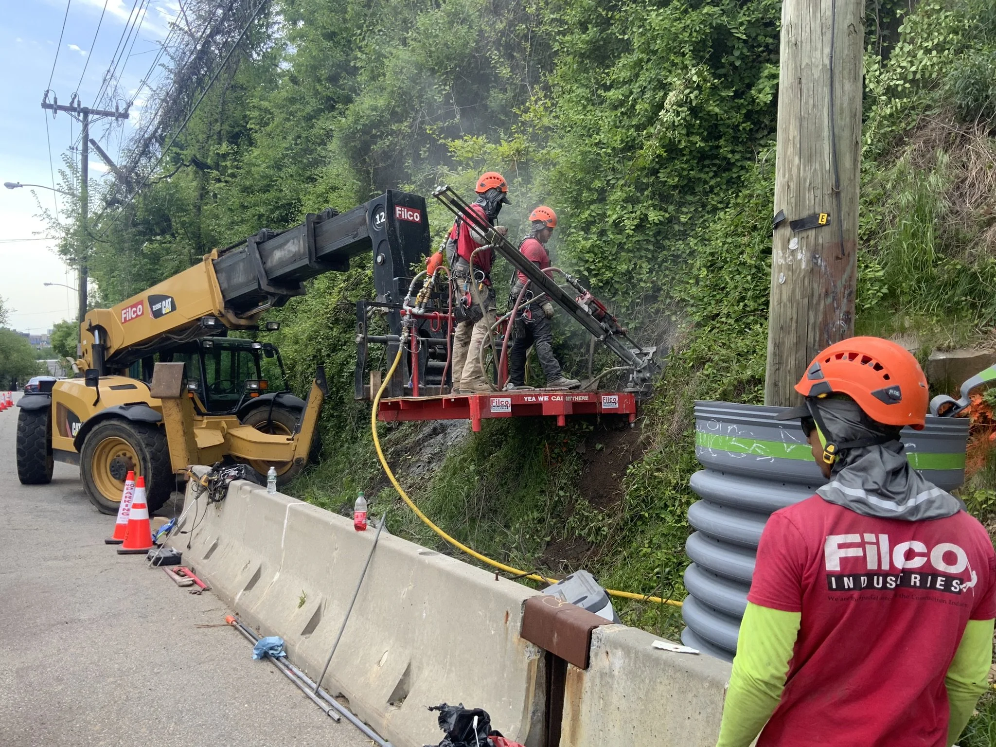 Workers in safety gear operating heavy machinery and tools along roadside to repair or maintain power lines on utility pole, with orange cones and barriers for safety.