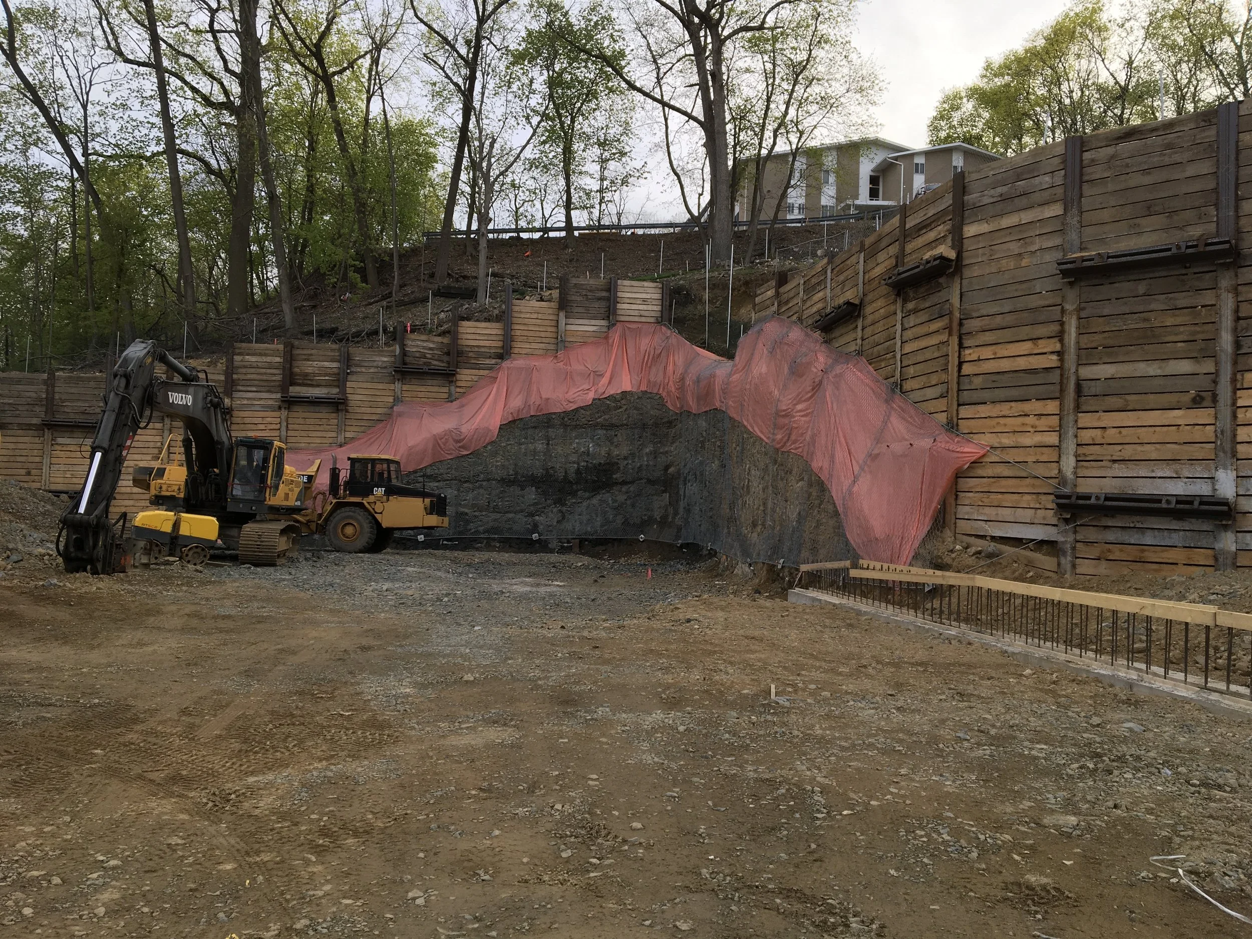 Construction site with excavator and bulldozer near retaining wall with protective netting, in a semi-wooded area.