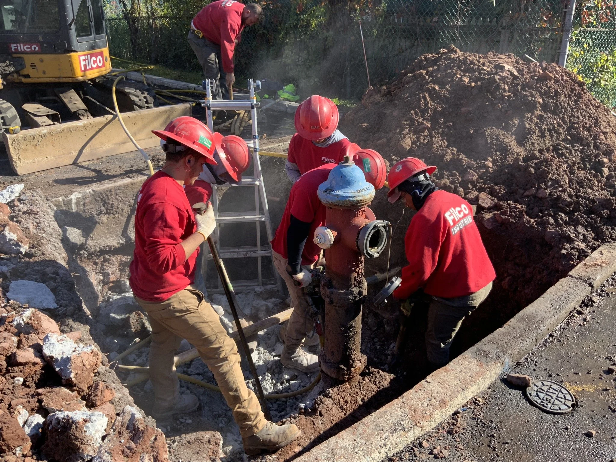 Construction workers wearing red shirts and helmets working on underground pipeline repair. They are using tools and equipment, surrounded by dirt and debris, with a large pile of dirt and construction machinery nearby.