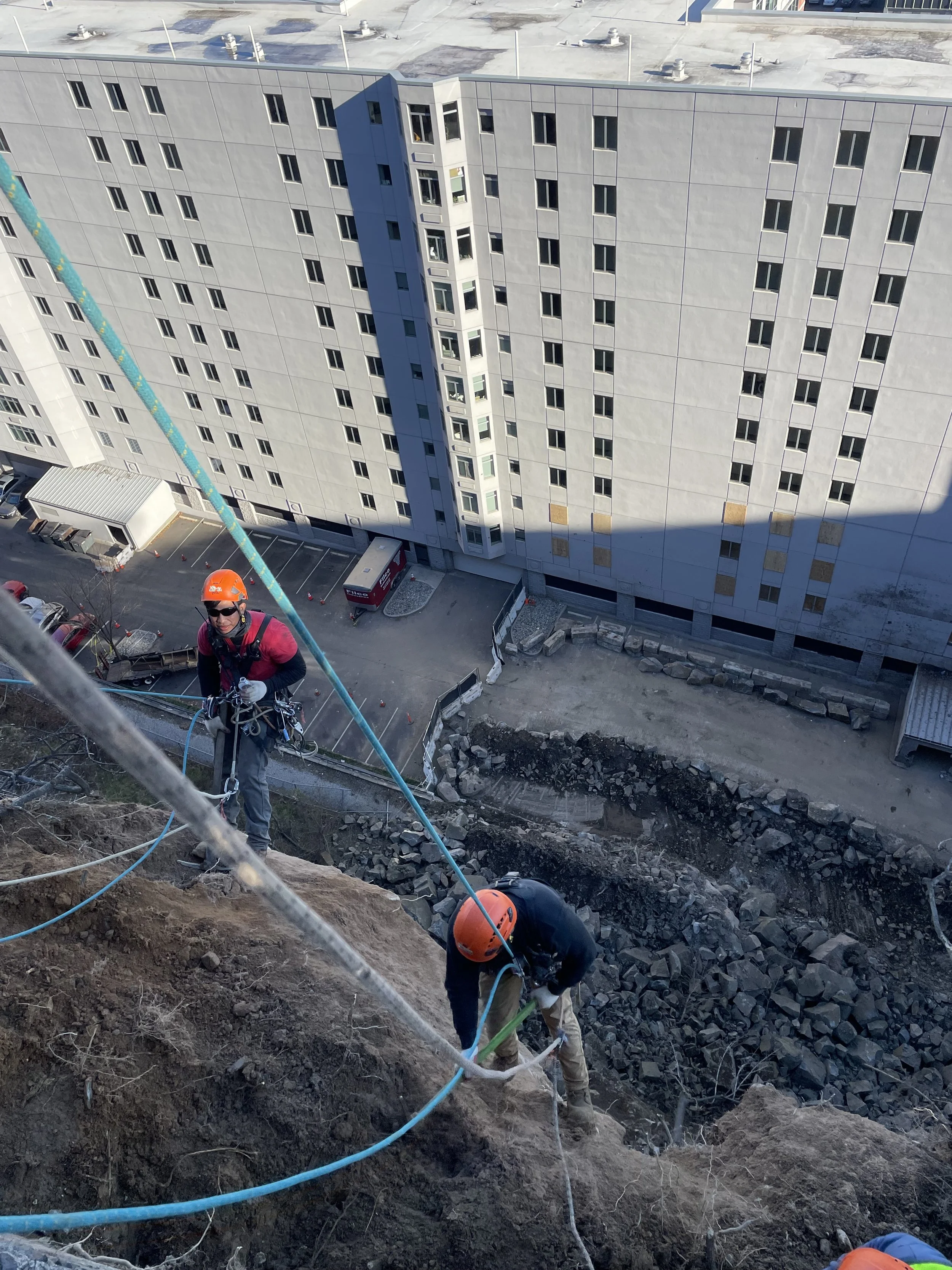 Two construction workers wearing orange helmets and harnesses are climbing a steep dirt and rock slope on a building site. One worker is using a green tool to secure the ropes, while the other is holding onto the ropes and looking down. A large multi