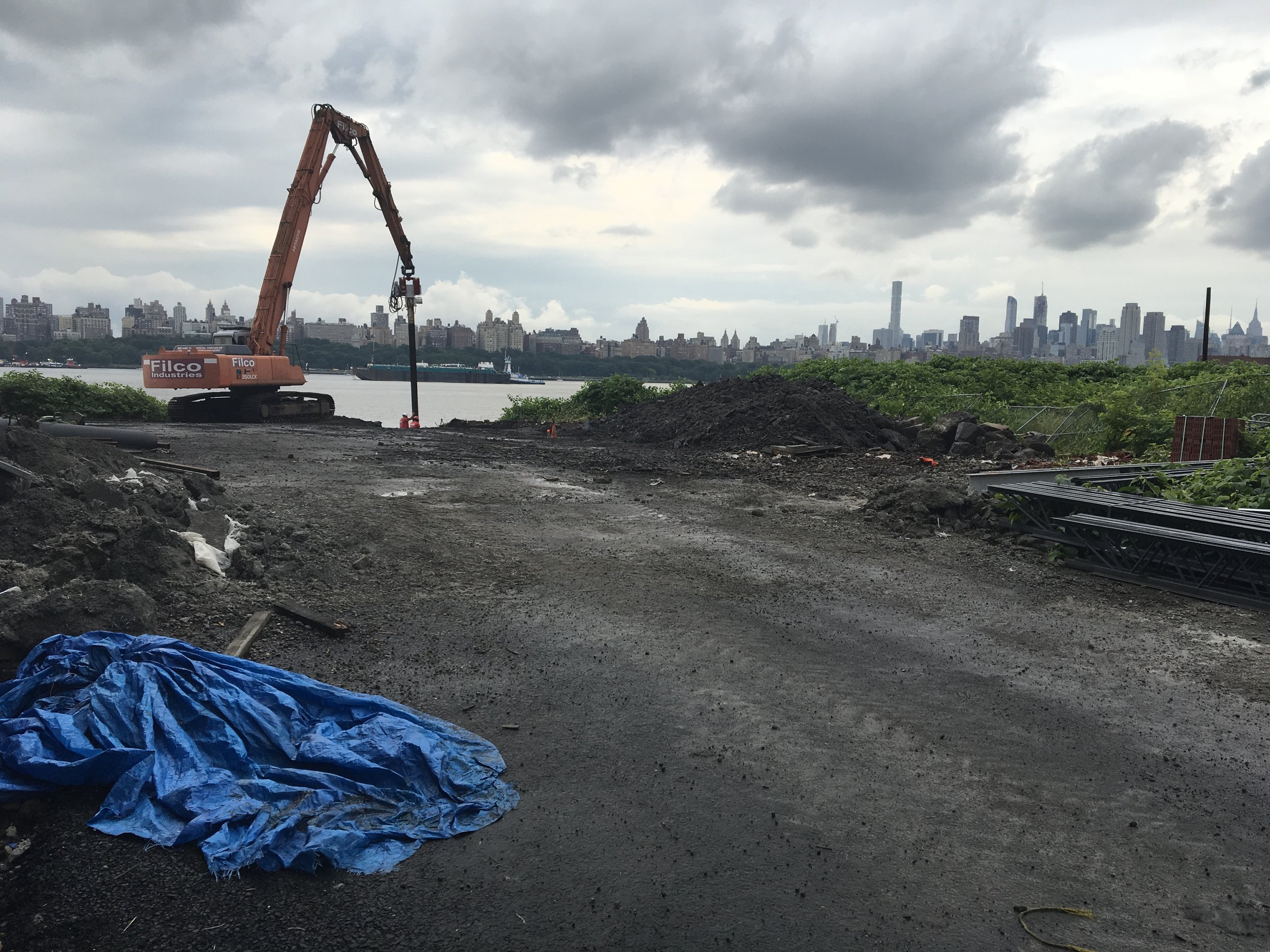 Construction site near water with an orange excavator and city skyline in the background under cloudy sky.