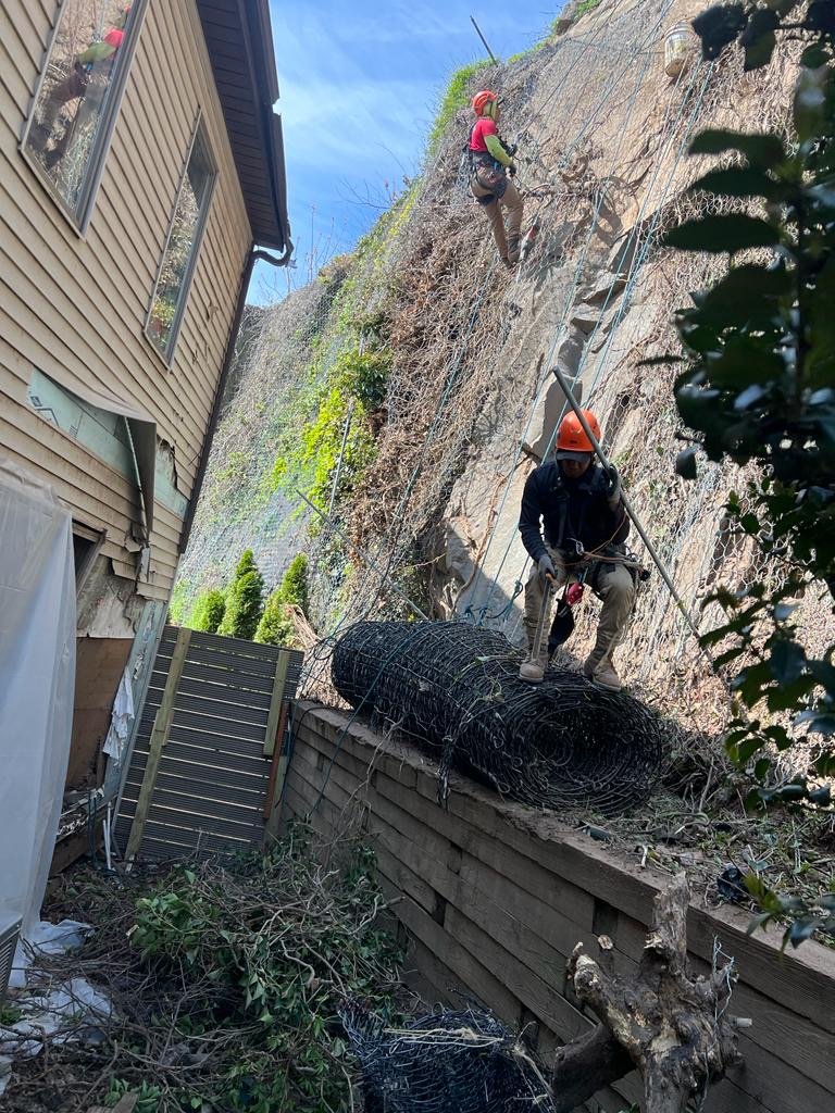 Two workers in safety gear climbing a steep outdoor rock face with the help of ropes and helmets, installing or repairing a wire fence.