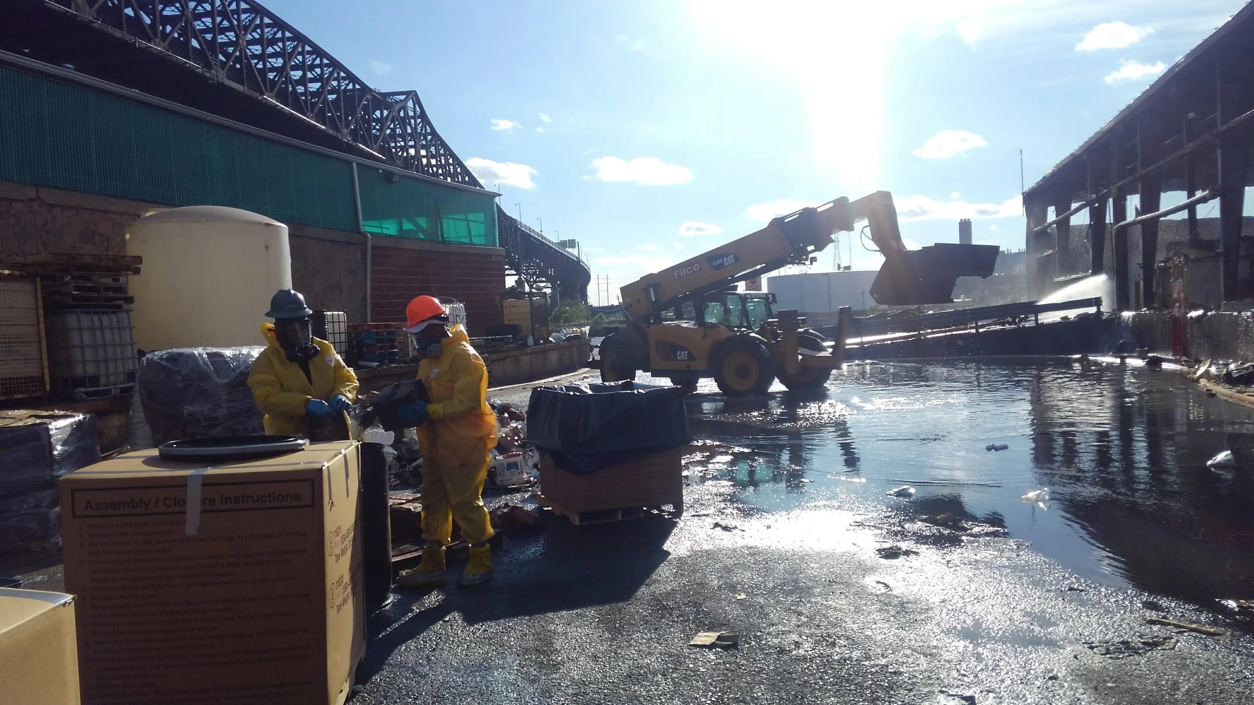 Workers wearing yellow protective suits, masks, and helmets at an industrial site with machinery, containers, and water. The scene appears to be that of cleanup or maintenance work near a building and a large overhead crane.