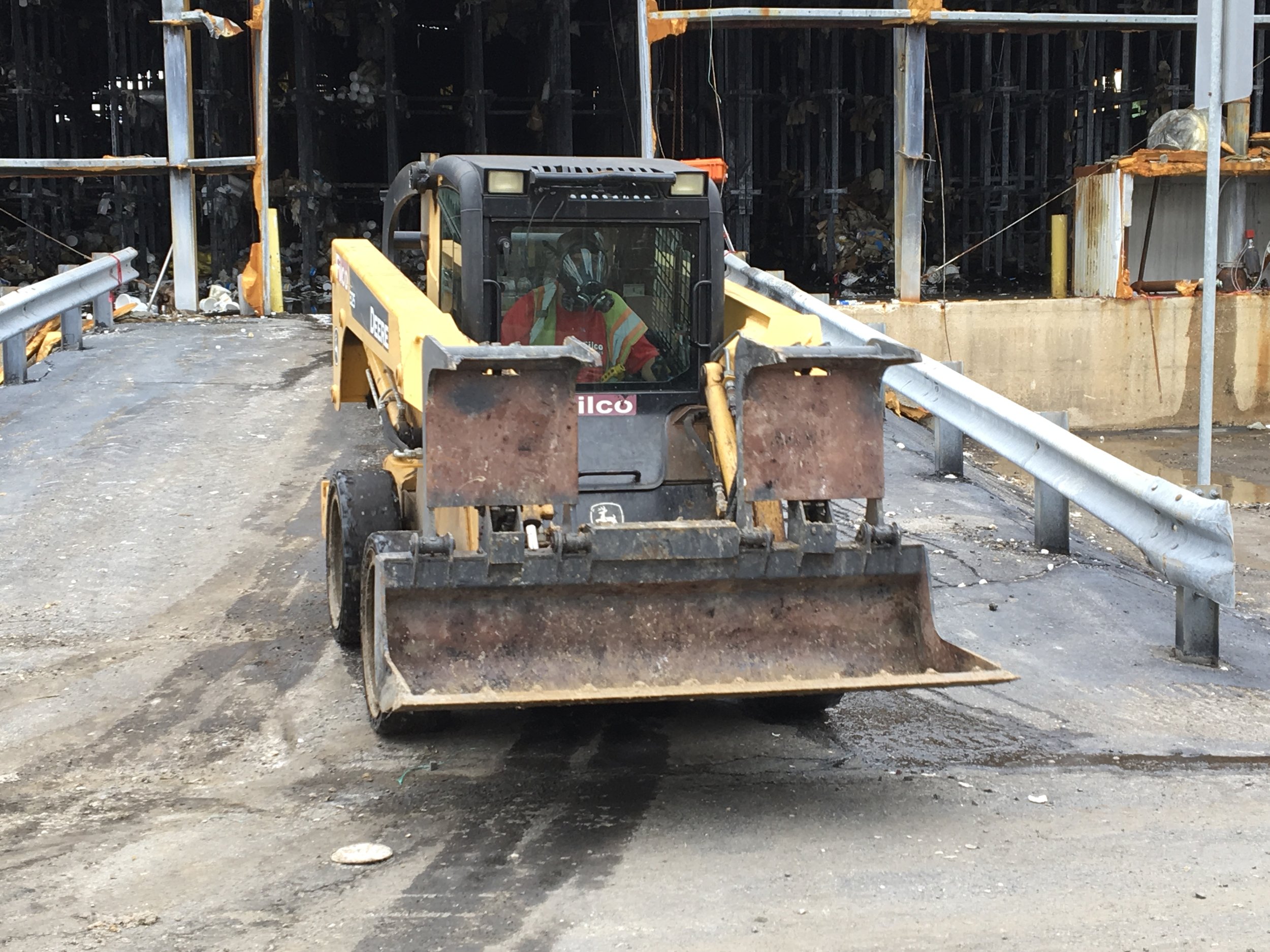 A construction worker operating a small John Deere bulldozer on a paved surface at a construction site, with steel guard rails and industrial storage in the background.