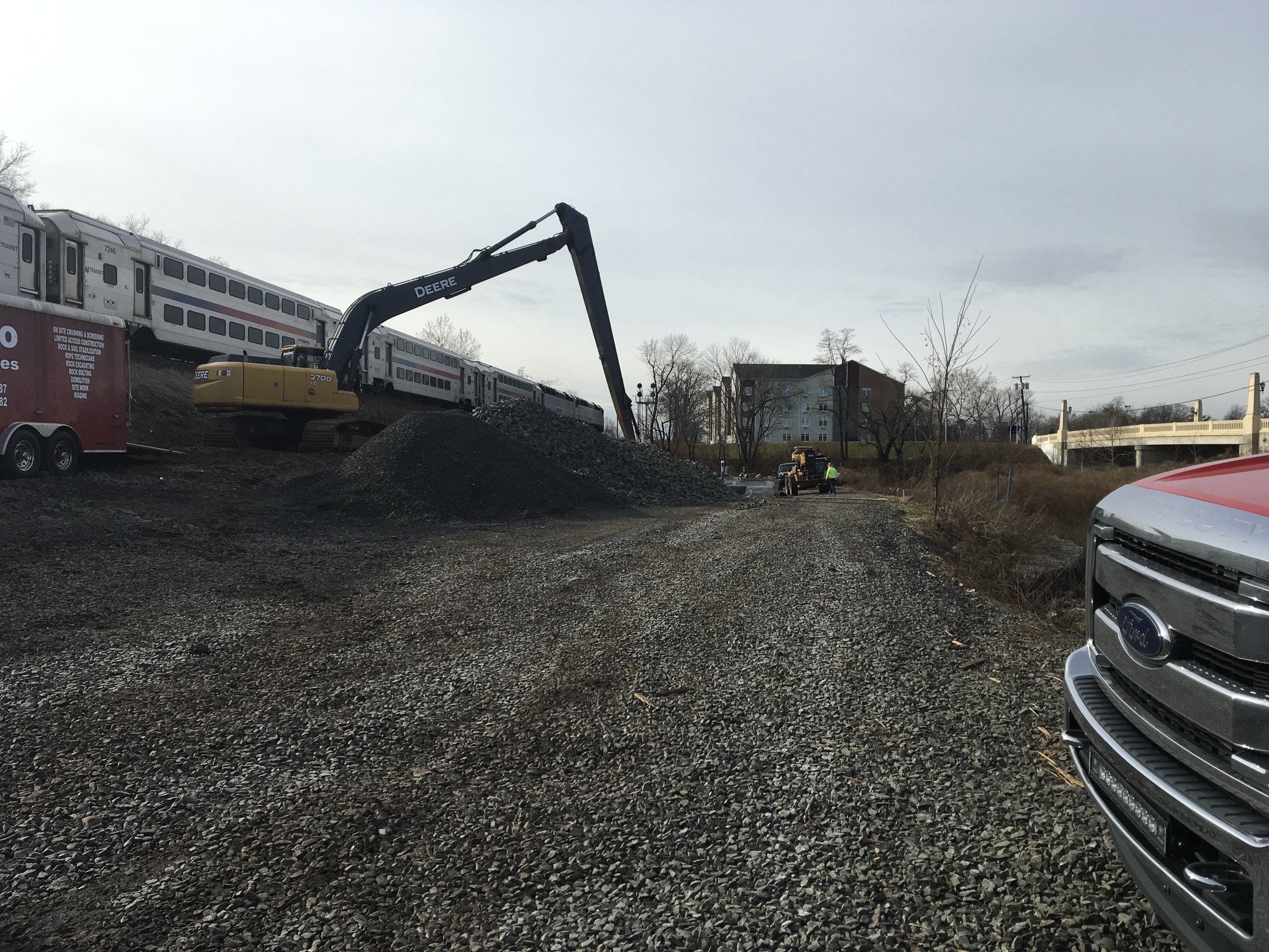 Construction site with a train on an elevated track, a yellow excavator digging into a pile of gravel, and a Ford vehicle partially visible in the foreground. Two workers are seen near a small construction vehicle in the background.