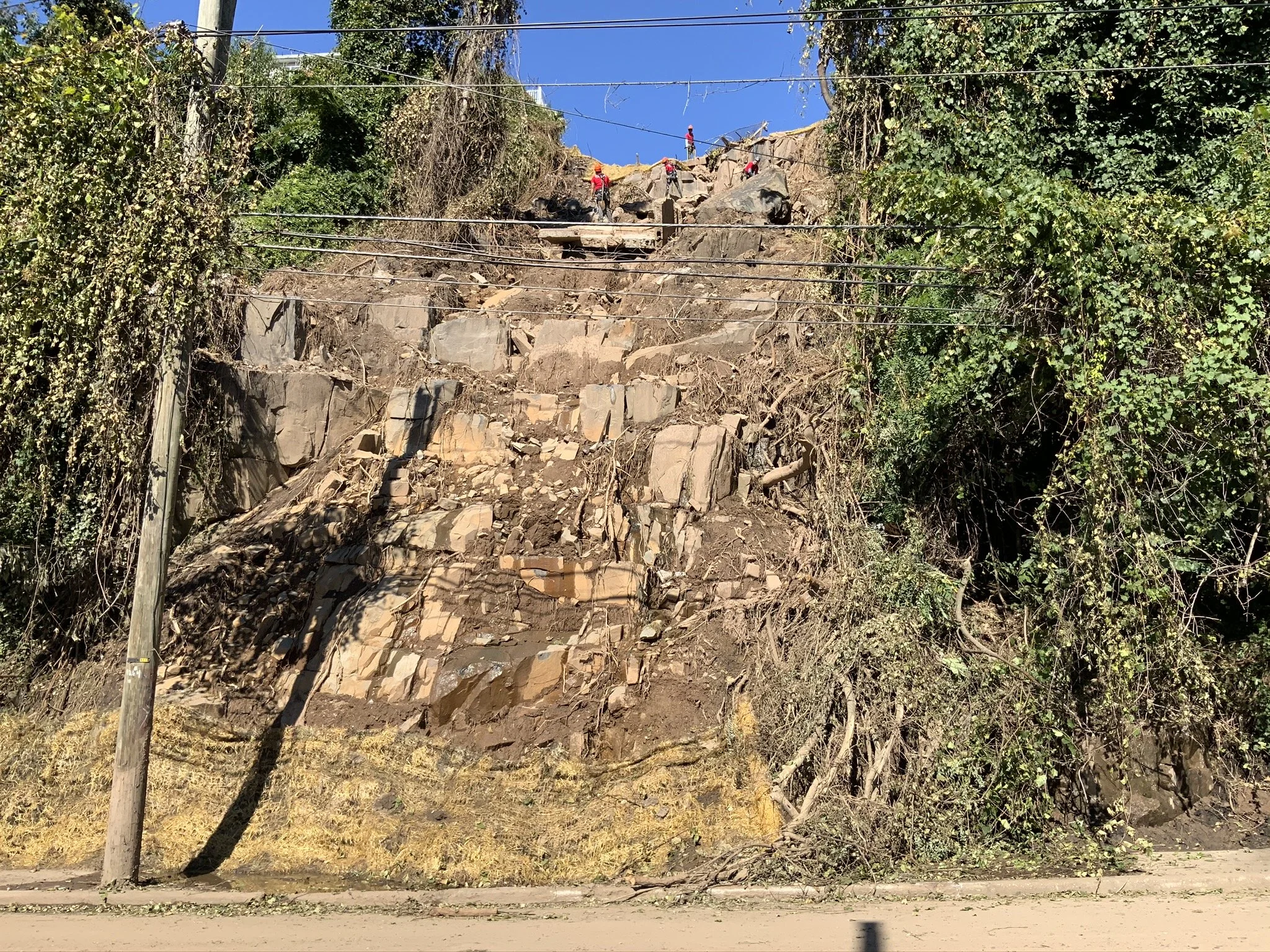 A hillside with rocks and dirt, partially cleared, with workers in red shirts working at the top, and utility wires across the scene, under a clear blue sky.