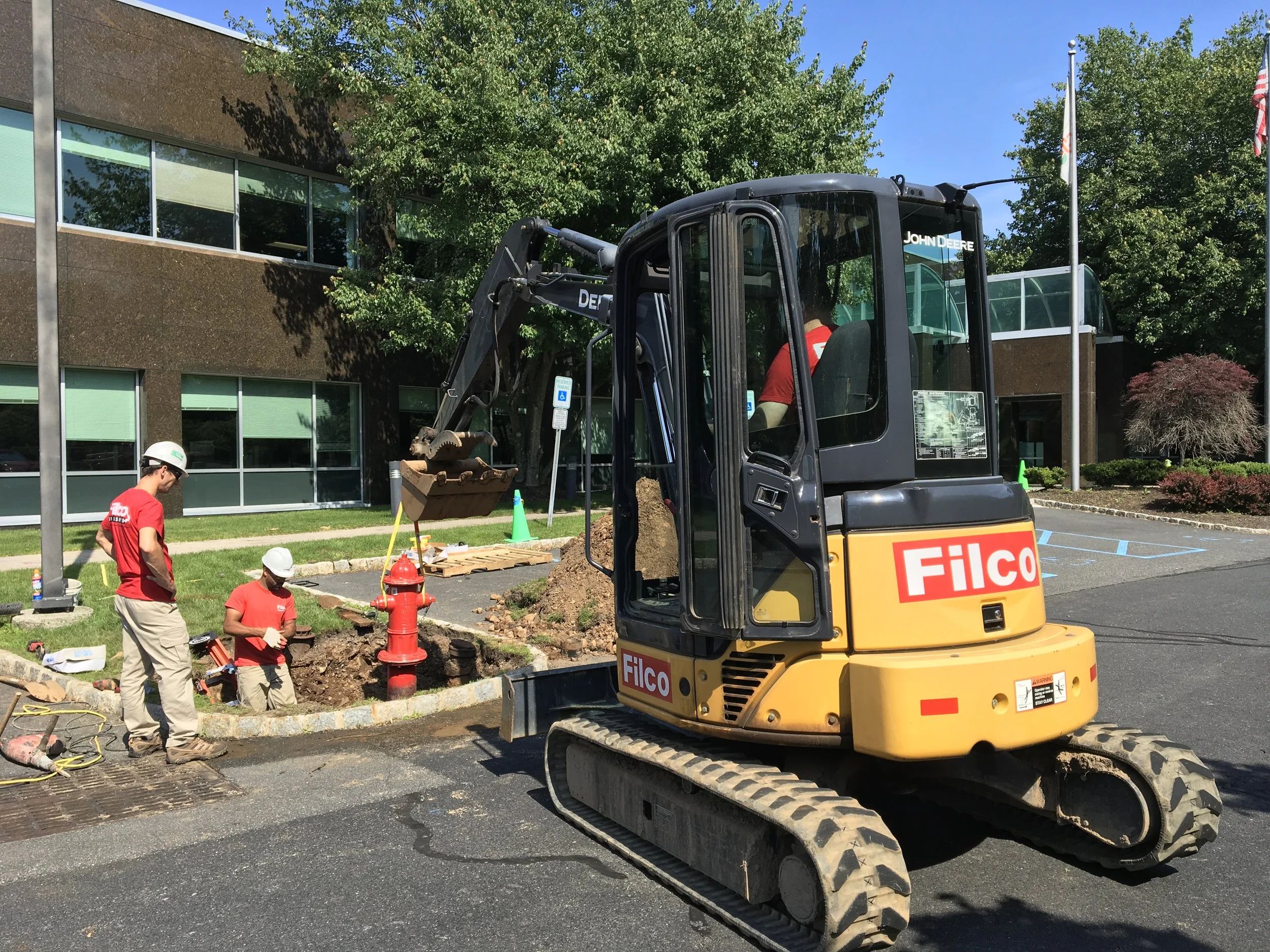 Construction workers in red shirts and white helmets working on a sidewalk with a small excavator labeled "Filco" nearby, in front of a modern office building with large windows and green trees.