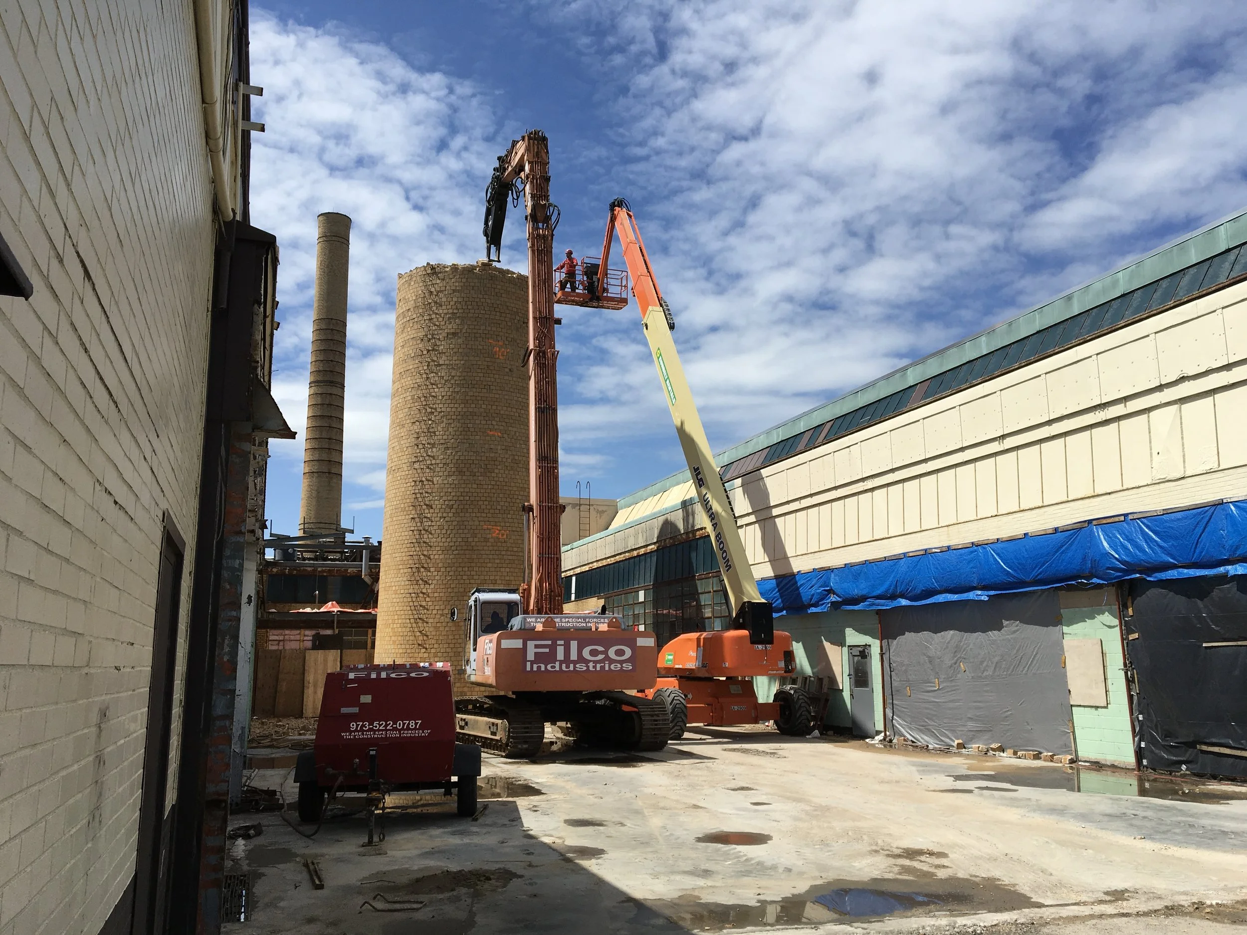 Construction site with a crane lifting equipment, with brick structures and an unfinished building on the right, under a partly cloudy sky.