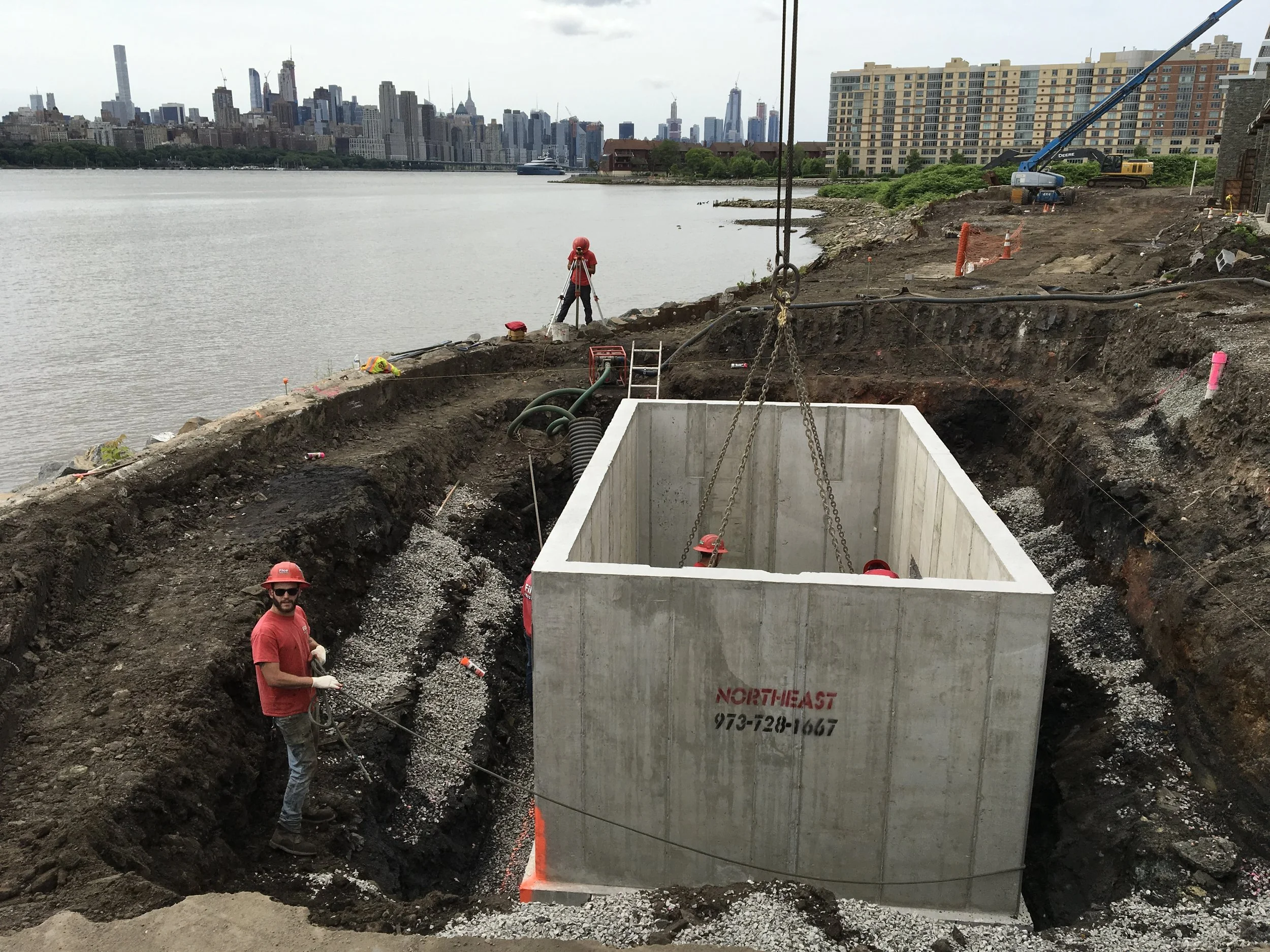 Construction workers installing a large concrete structure in a trench along a riverside with cityscape in the background.