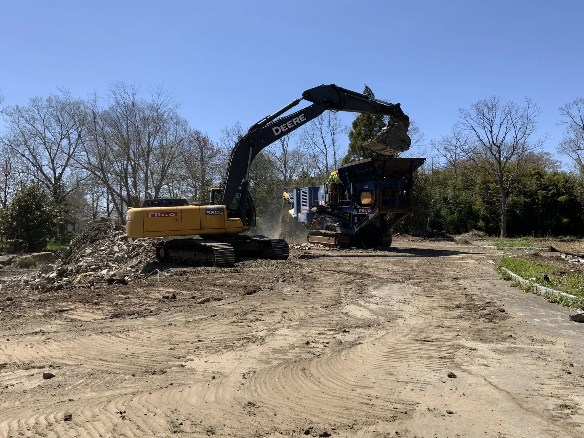 Construction site where an excavator and a wood chipper are working together, with workers nearby, dirt and trees in the background, and a clear blue sky.