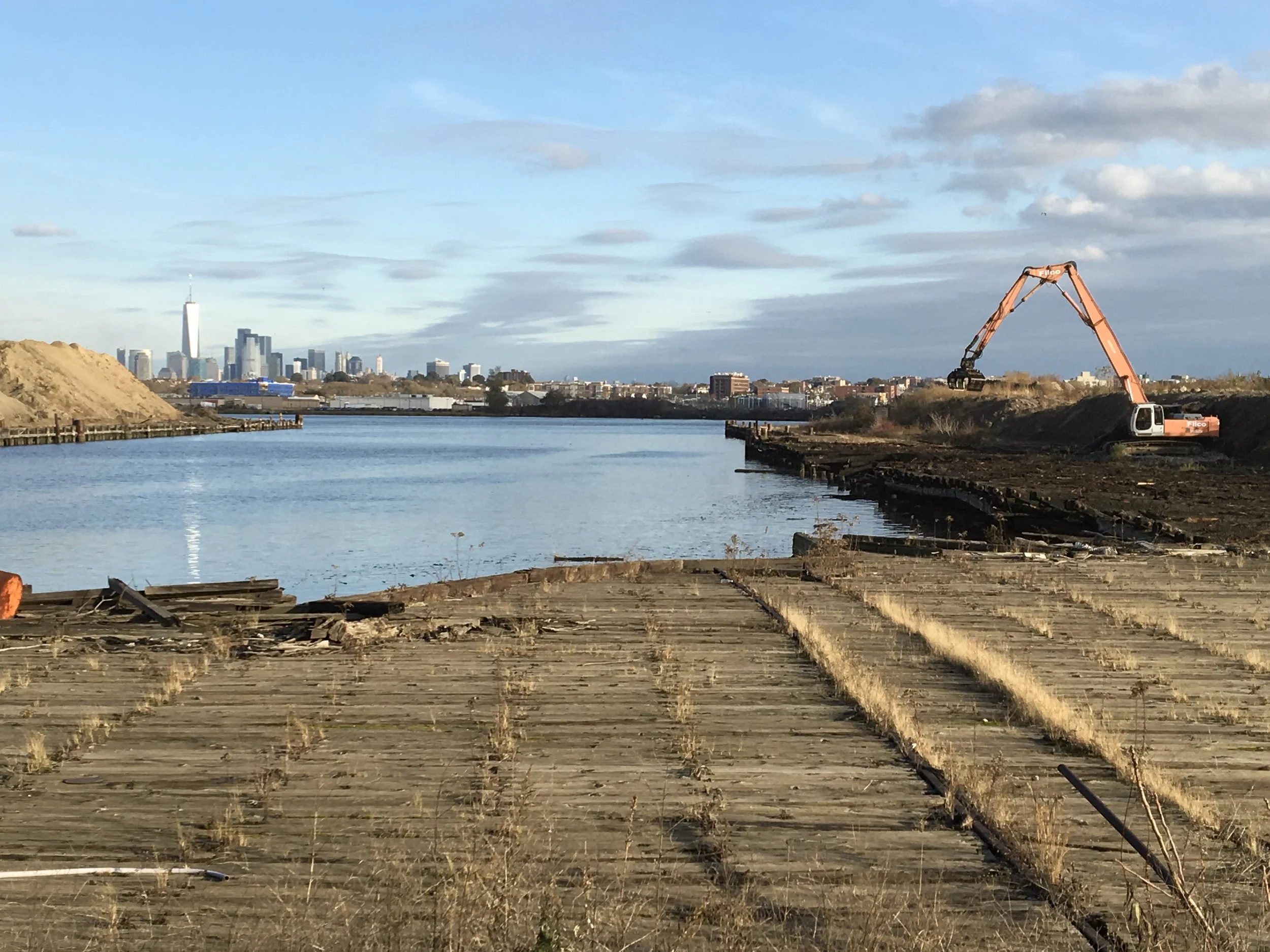 Construction site along a river with an excavator working on the shoreline, city skyline in the background, blue sky with scattered clouds.