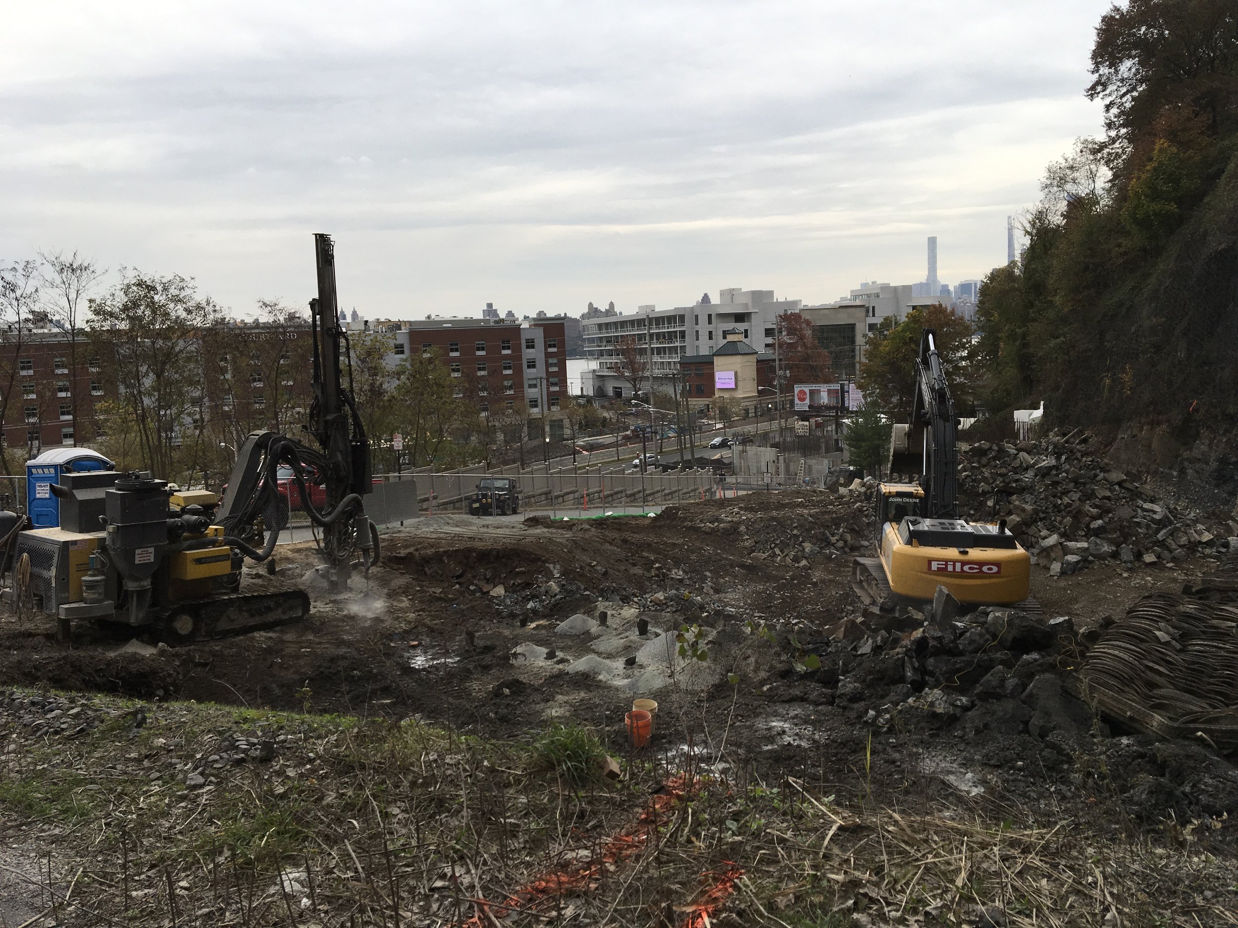 Construction site with two excavators working on dirt and rocks, surrounded by urban buildings and a hill with trees.