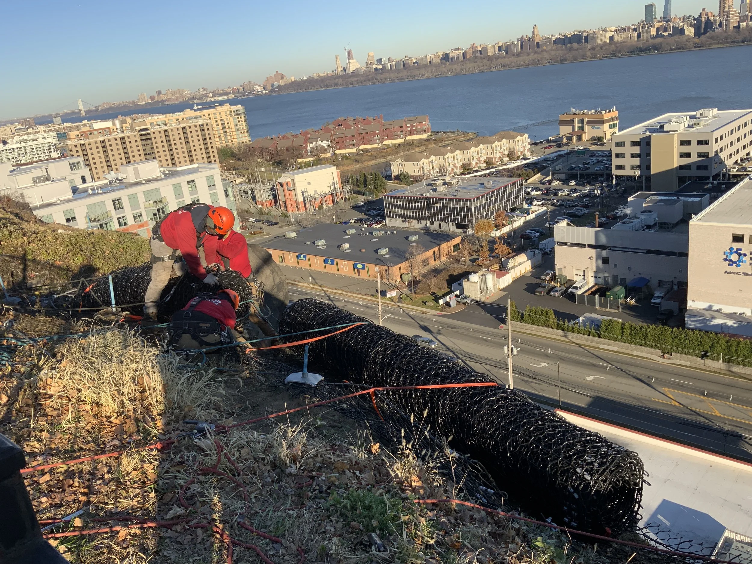 Workers installing or inspecting a black mesh safety barrier on a hillside overlooking a city skyline and water.