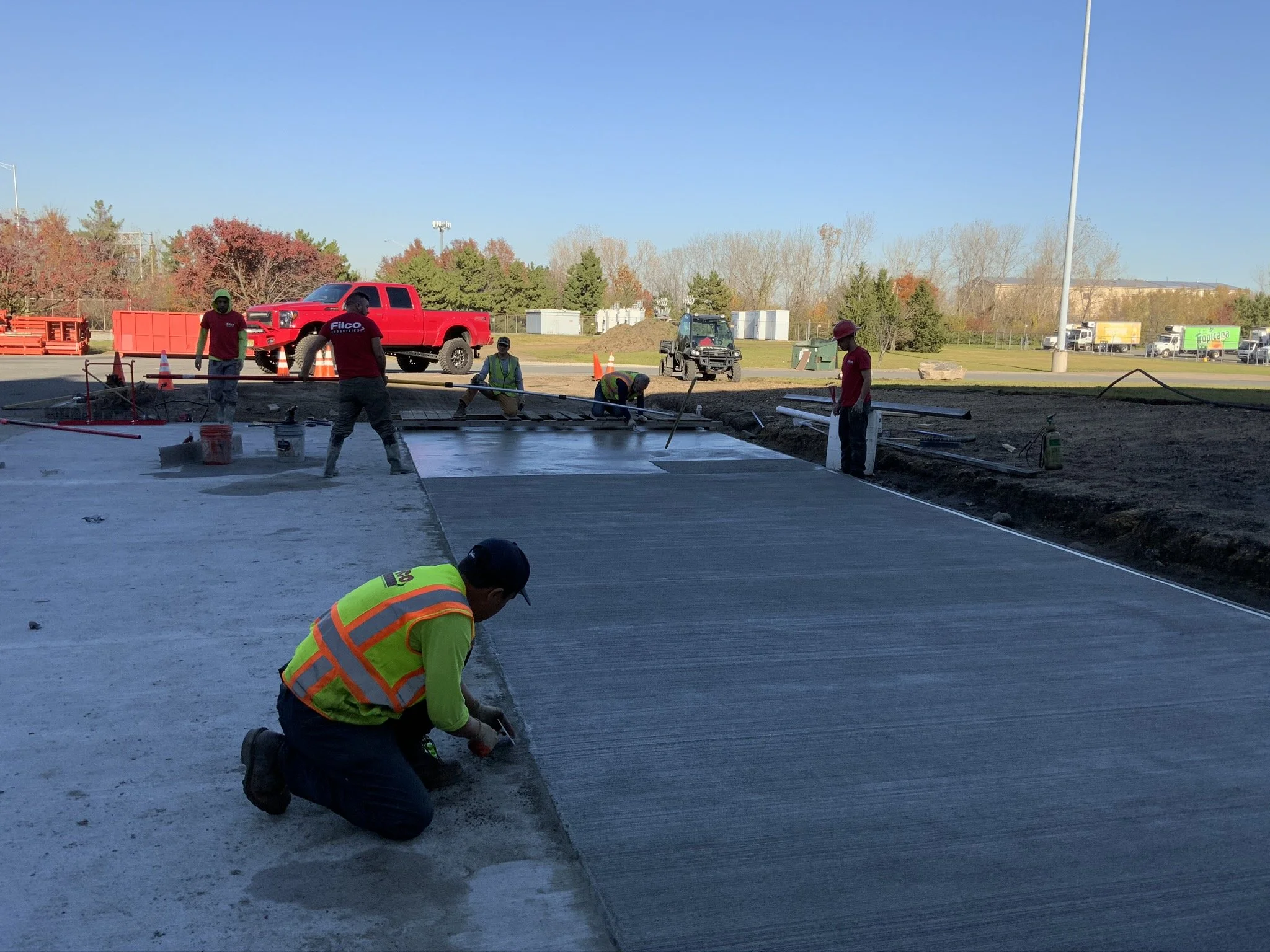 Construction workers are pouring and leveling concrete on a large outdoor site, with trucks and construction equipment in the background.