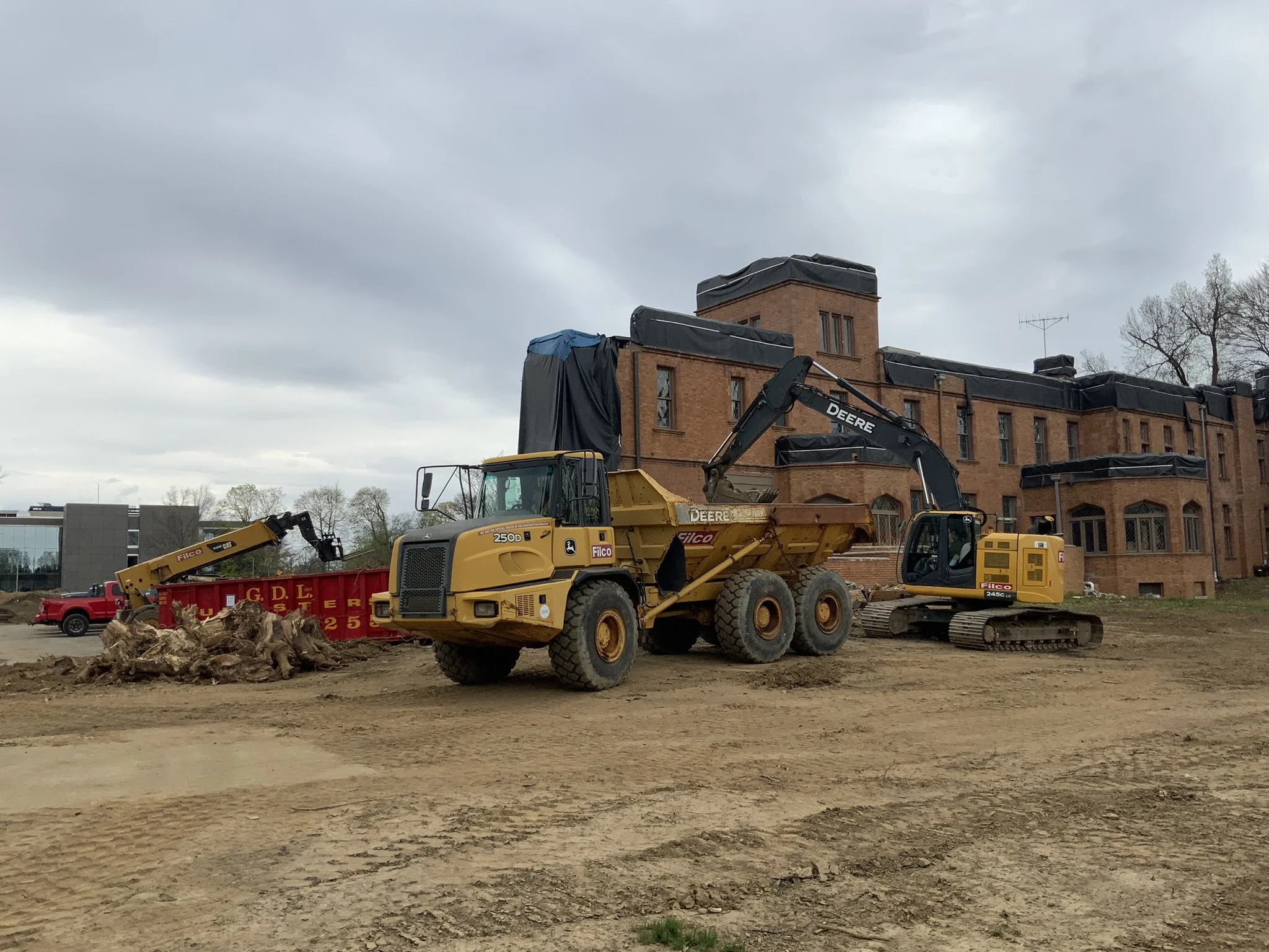 Construction site with a yellow dump truck, a small bulldozer, and a small excavator working on ground in front of a large brick building under cloudy sky.