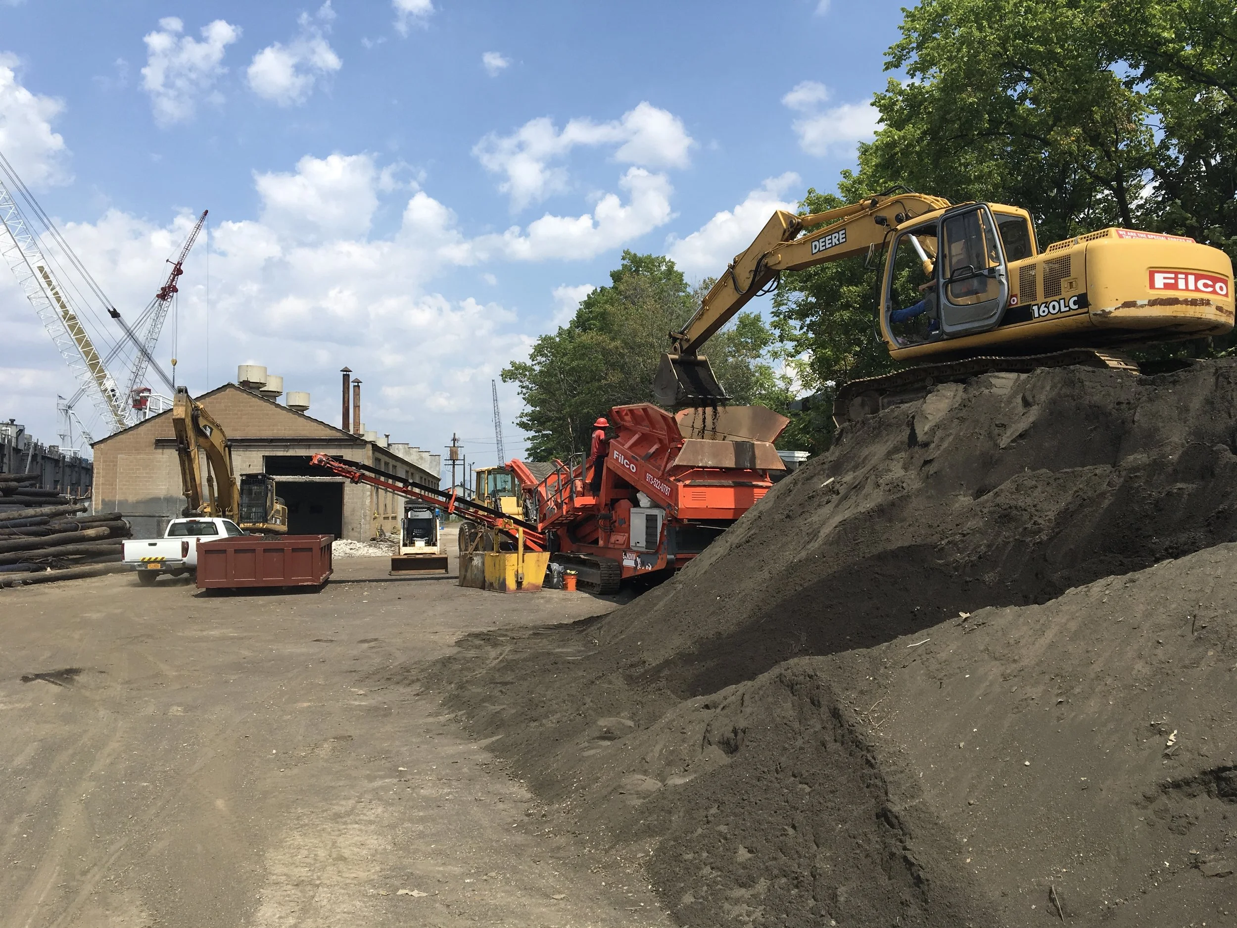 Construction site with excavators, bulldozer, and building in background, dirt and soil in foreground, blue sky with clouds above.