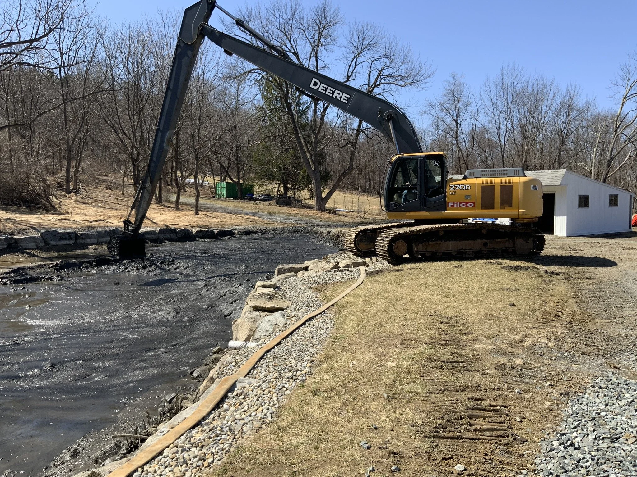 A large excavator is operating by a river, with a small white building and trees in the background, during the daytime.