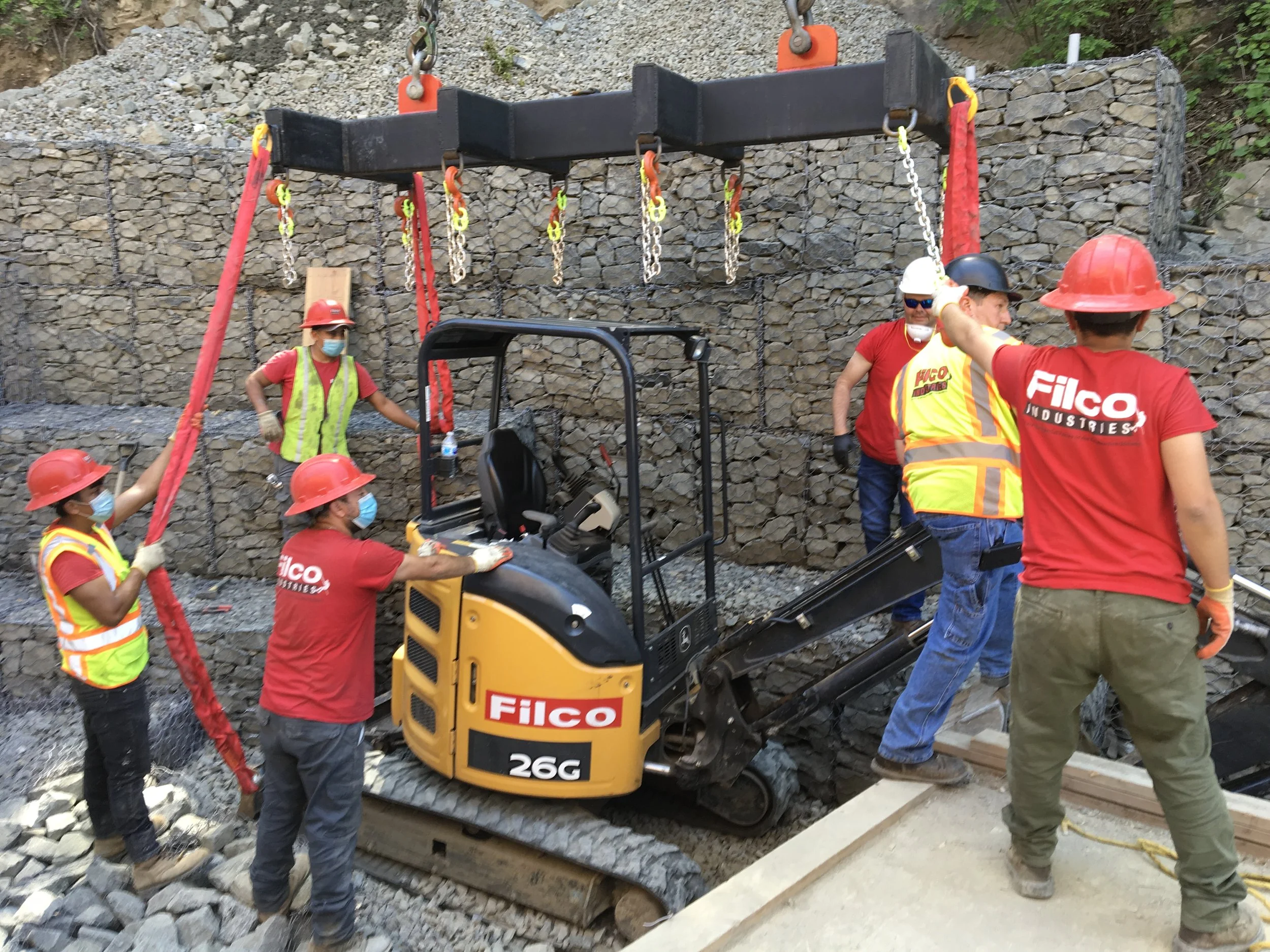 Construction workers operating a small excavator and installing a retaining wall with rocks, all wearing safety gear including helmets and masks.