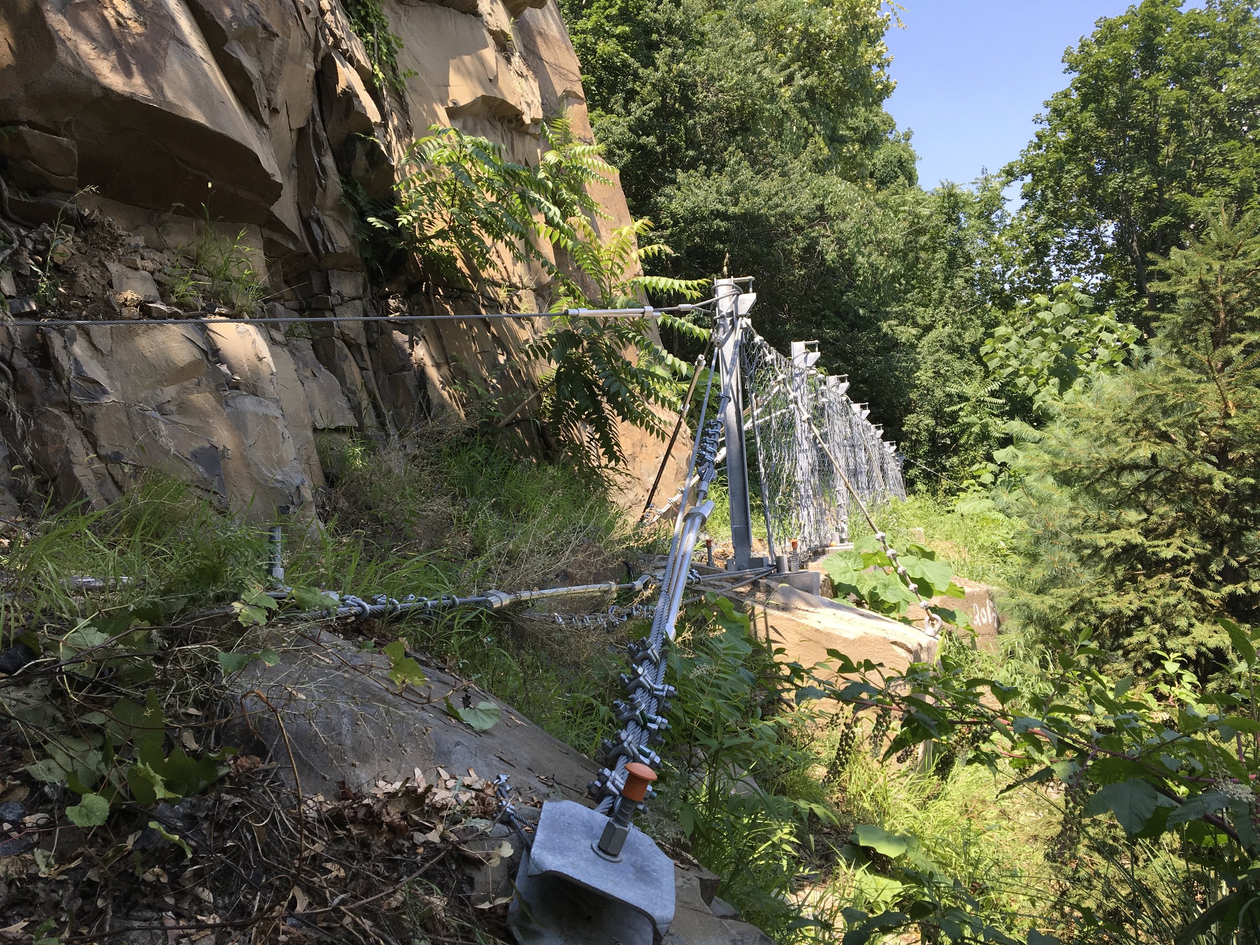 A set of safety cables and anchors installed along a rocky hillside with green trees and blue sky in the background.