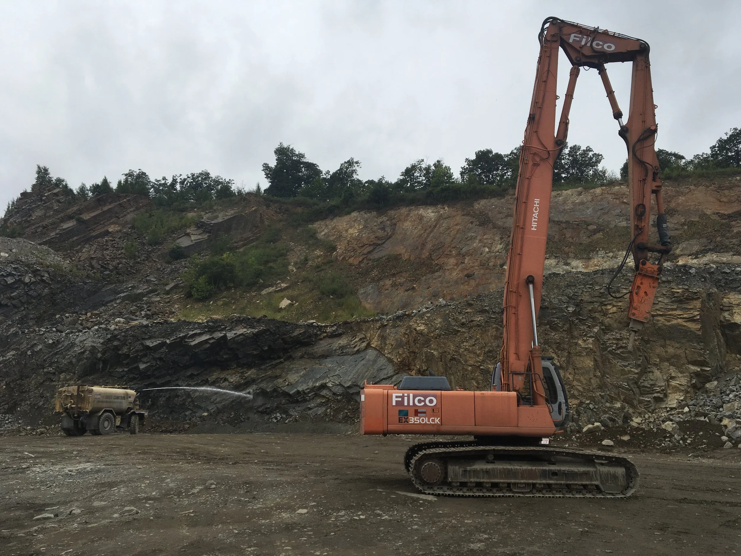 An orange excavator with tracks working on a rocky site with an overcast sky, and a truck spraying water in the background.