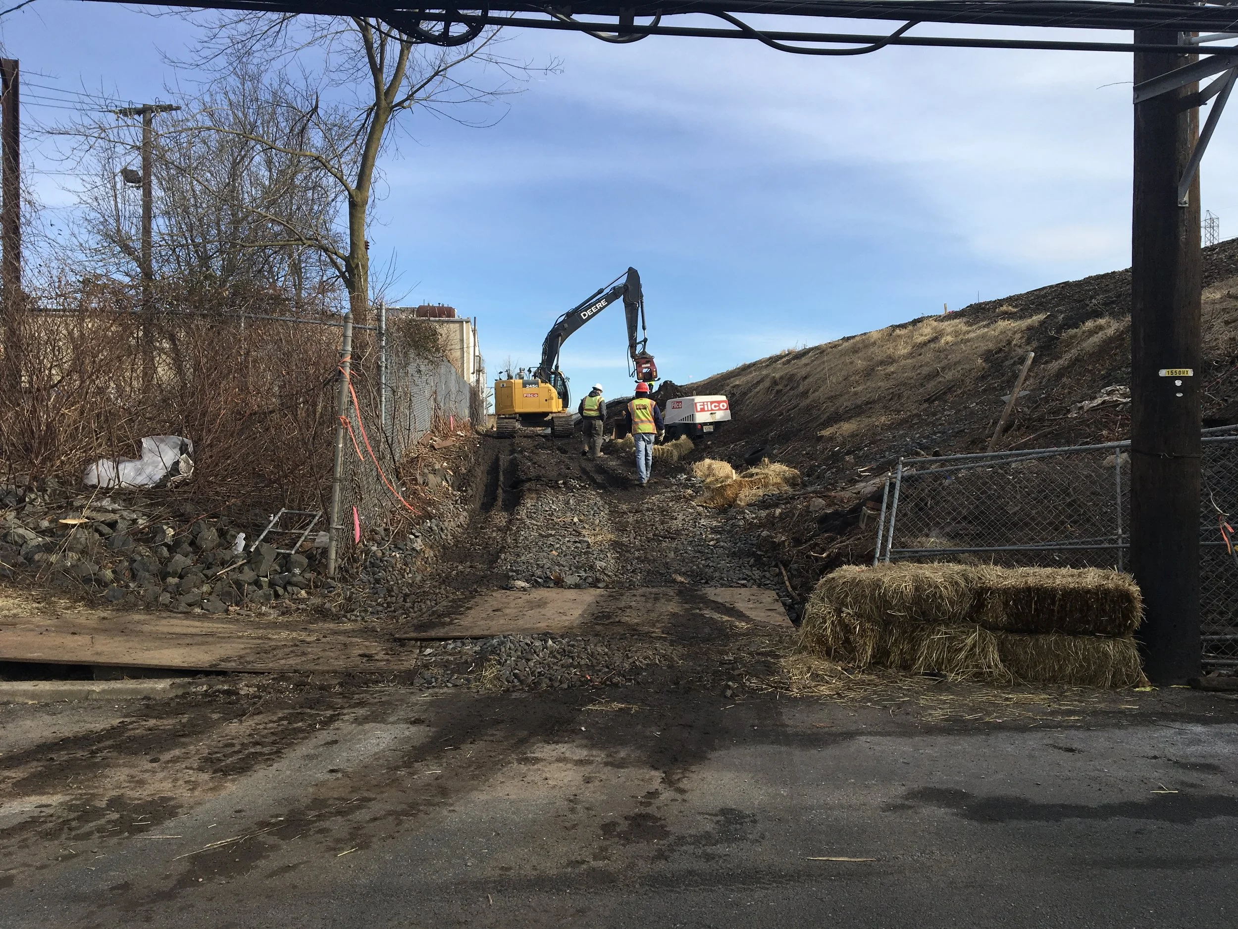 Construction workers operating an excavator on a dirt and gravel pathway, with hay bales and fencing along the sides, under a clear blue sky.