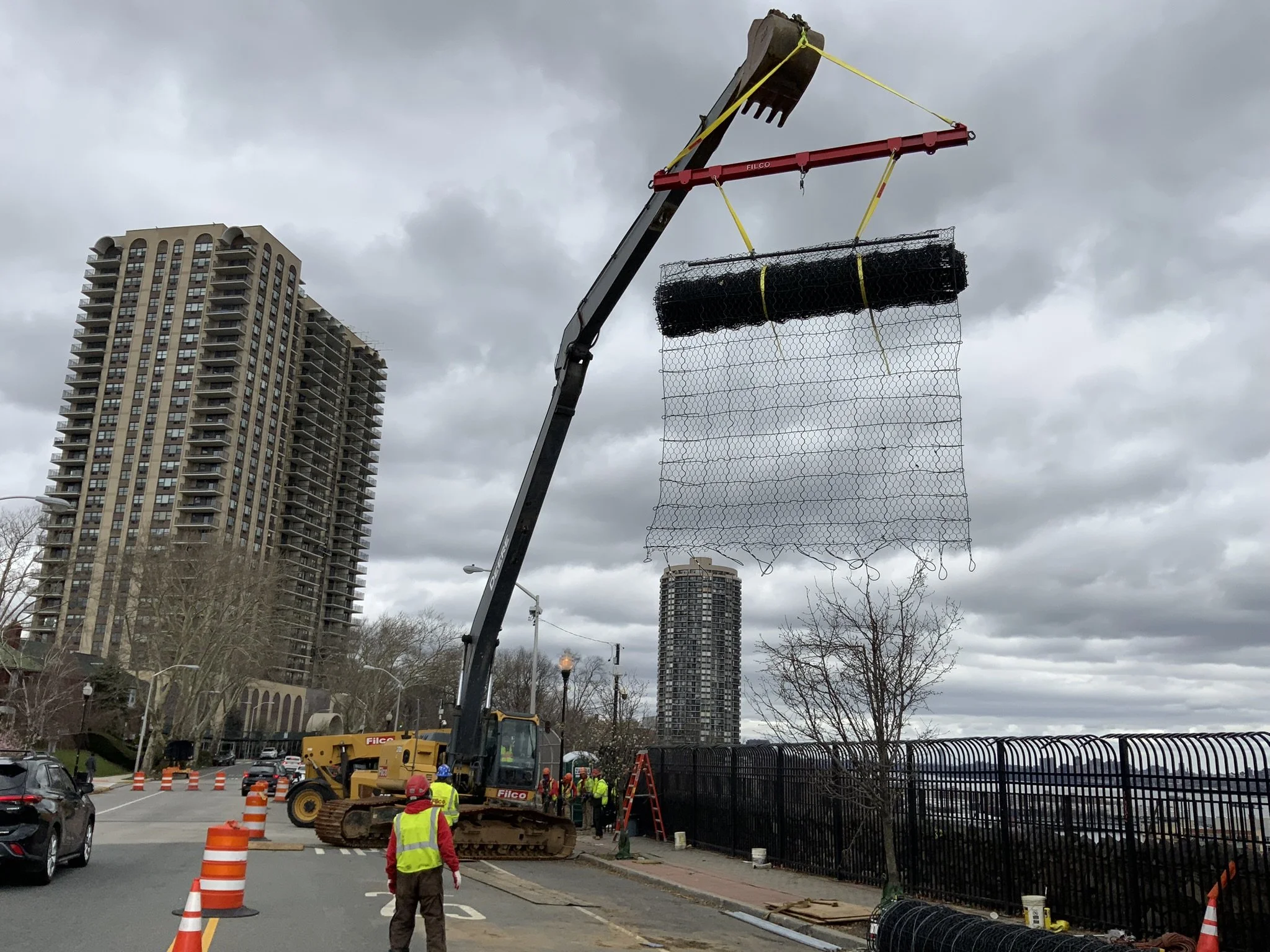 Construction workers operating a crane to install a black wire mesh fence on a sidewalk, with high-rise buildings and cloudy sky in the background.