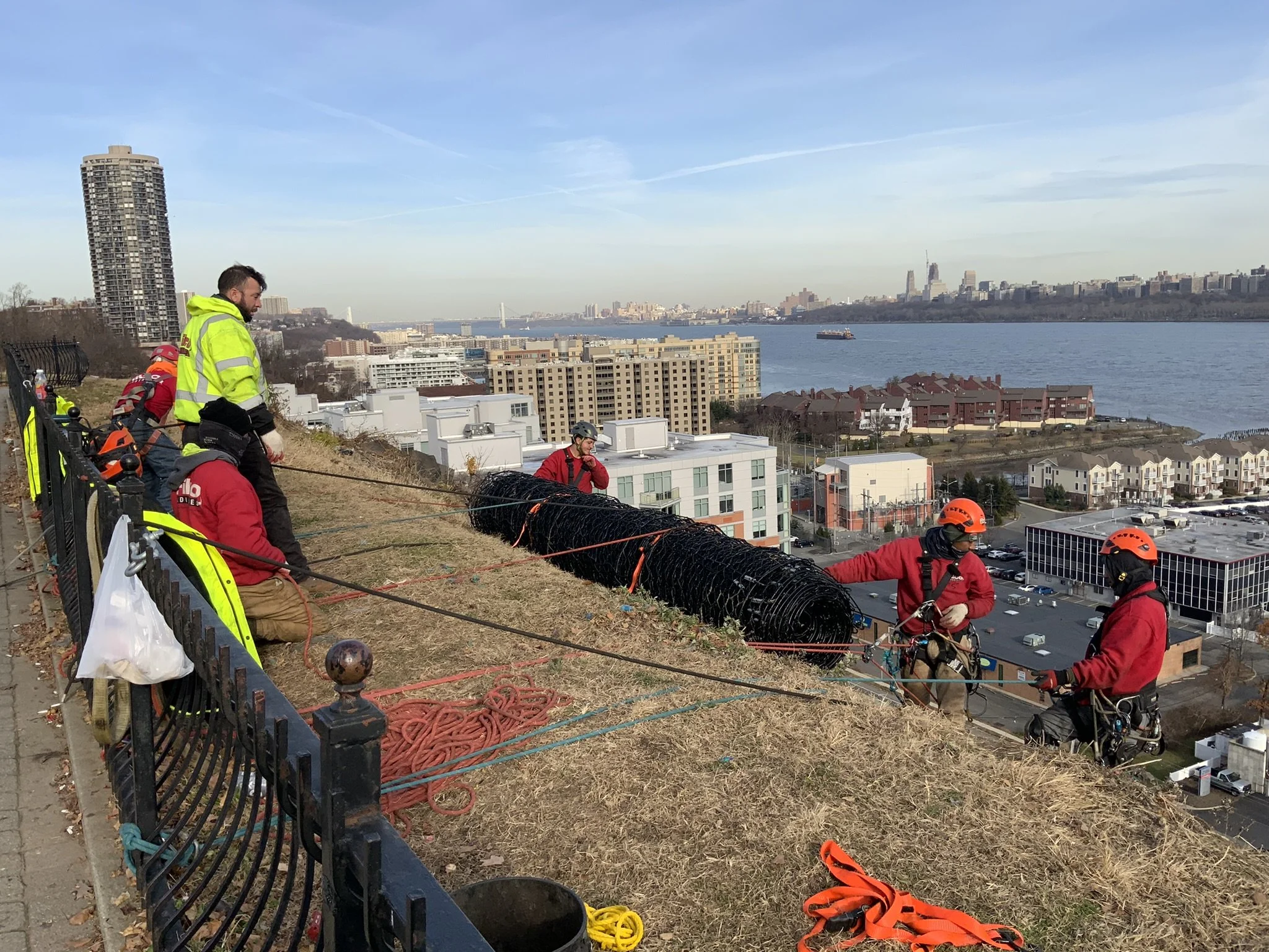 Group of workers on a hillside repair or install a large black barbed wire security fence with a cityscape and river in the background.