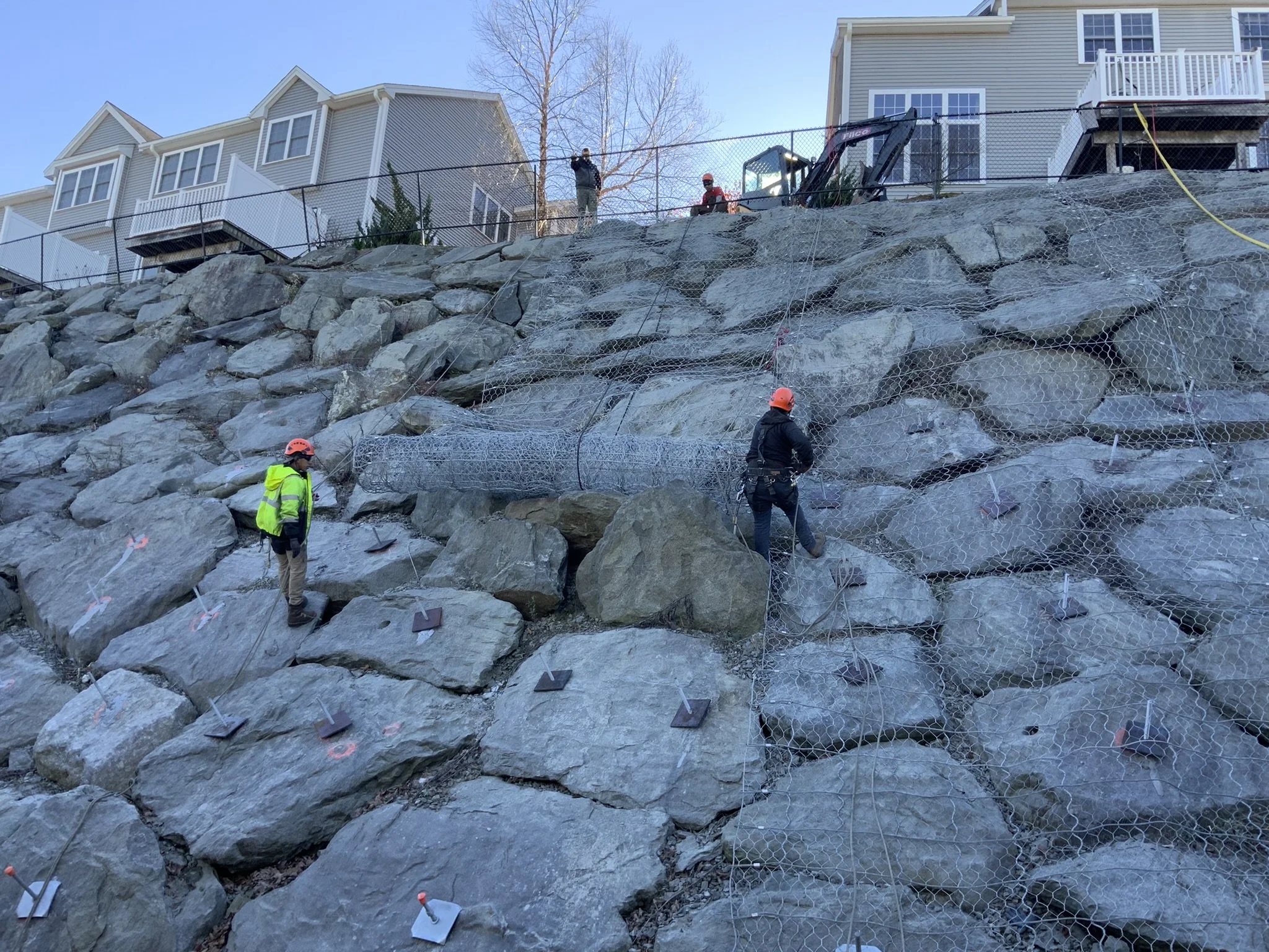 Workers in hard hats installing rock reinforcement mesh on a rocky hillside next to residential houses.