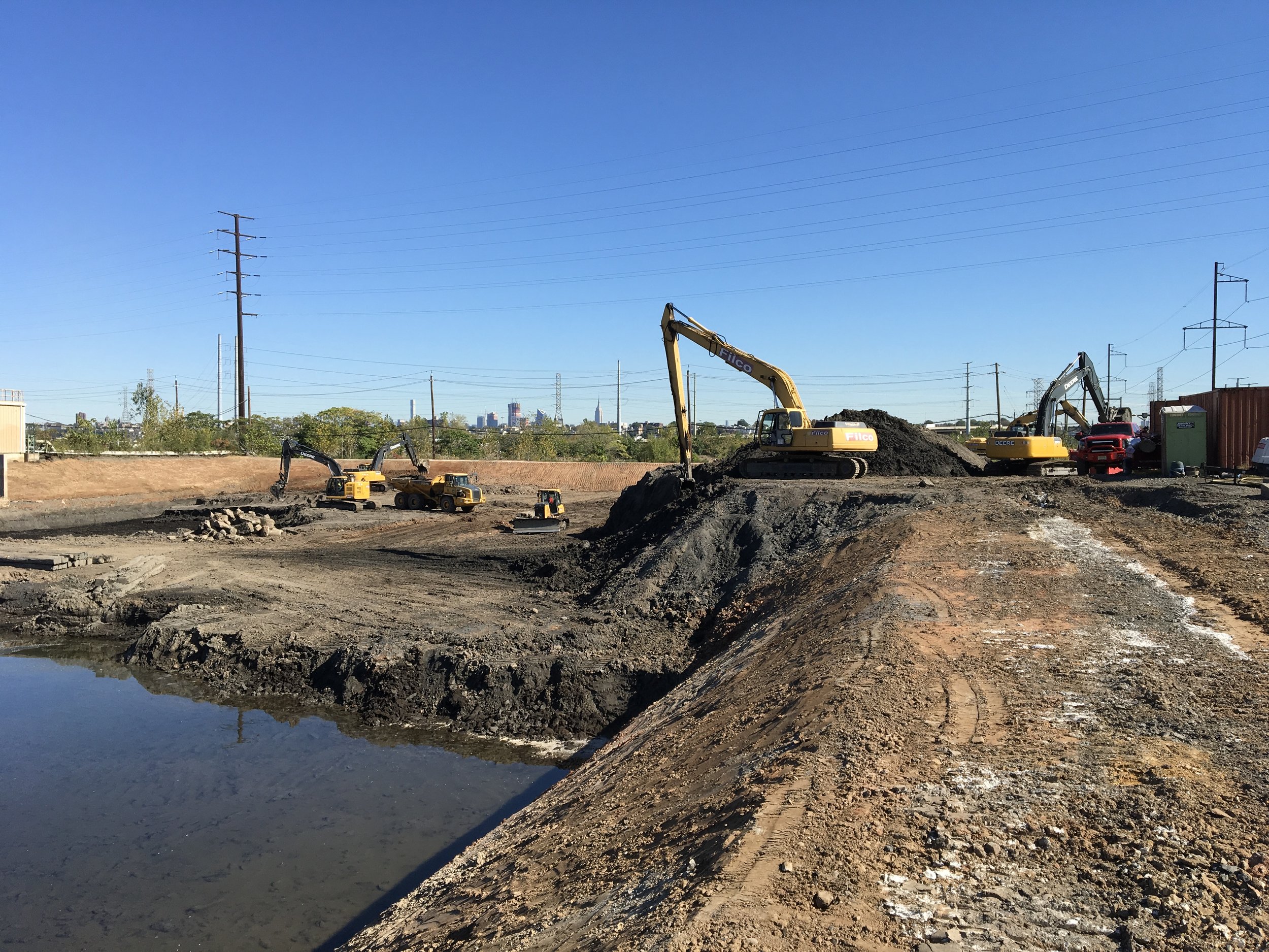 Construction site with excavators working on the dirt, and a body of water in the foreground. Clear blue sky and city skyline in the distance.