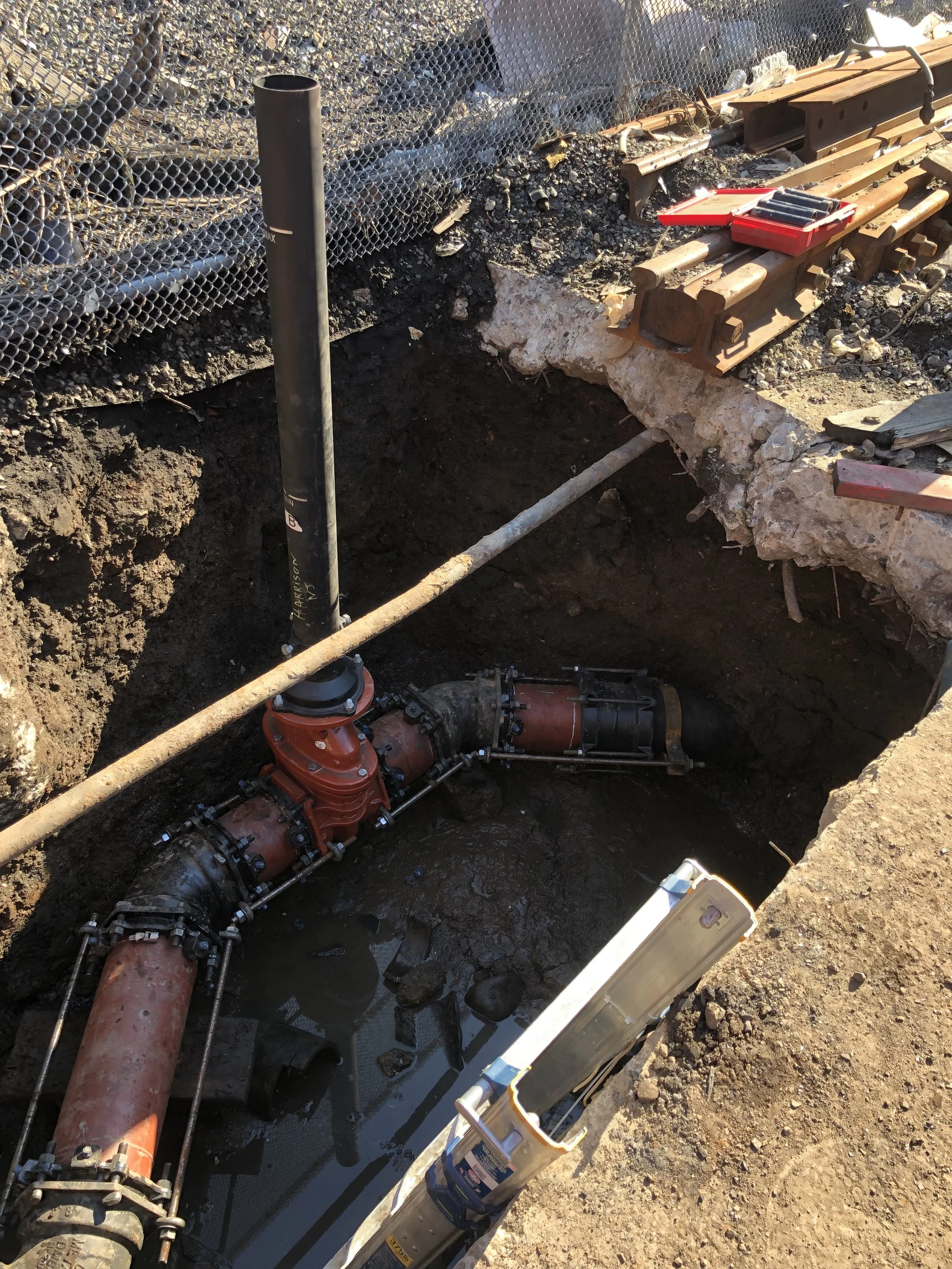 Underground plumbing with large pipes and a valve in a construction trench, surrounded by dirt and construction materials.