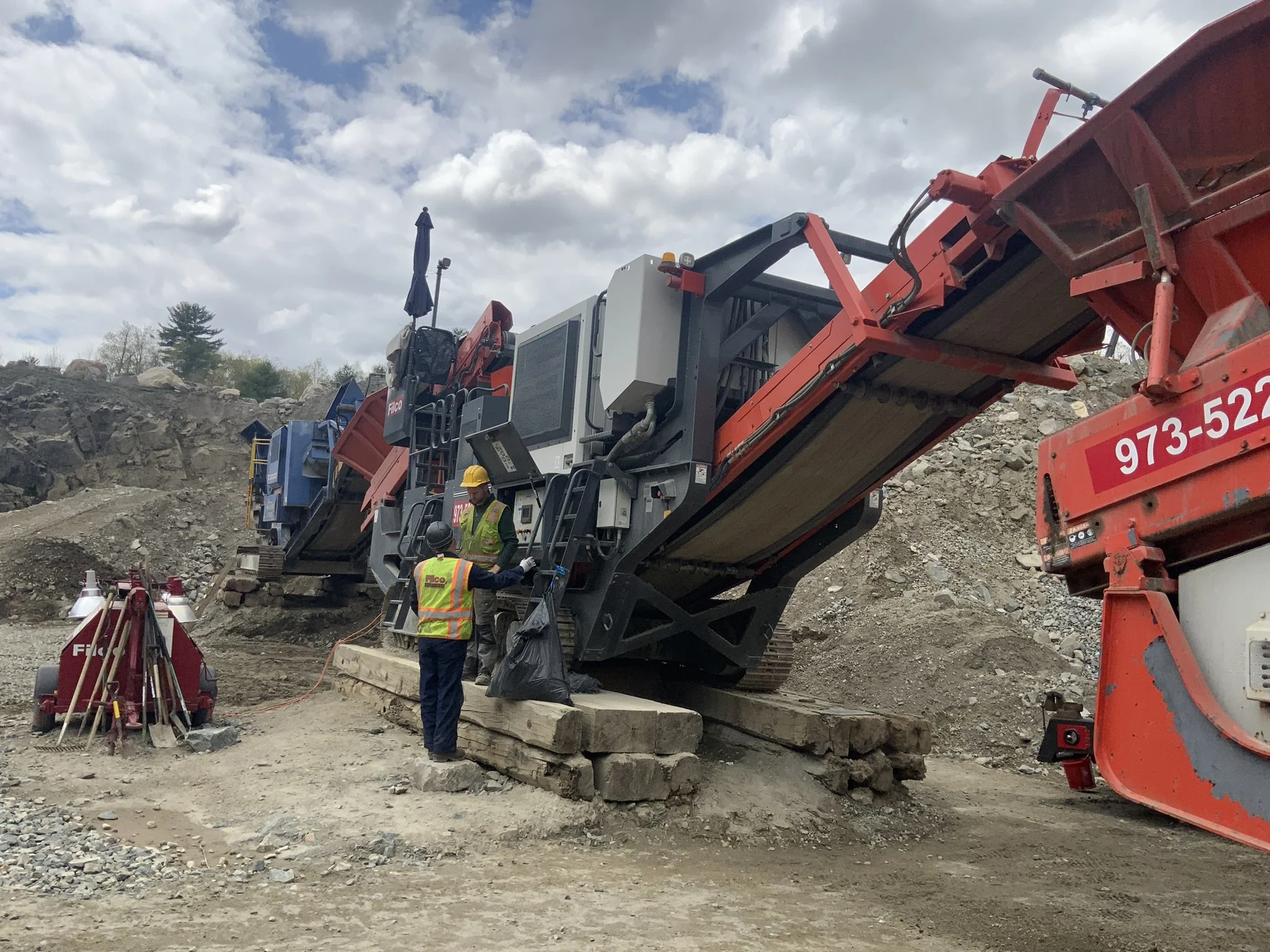 Construction workers standing next to a large piece of construction equipment on a dirt site, with rocky terrain and a cloudy sky in the background.